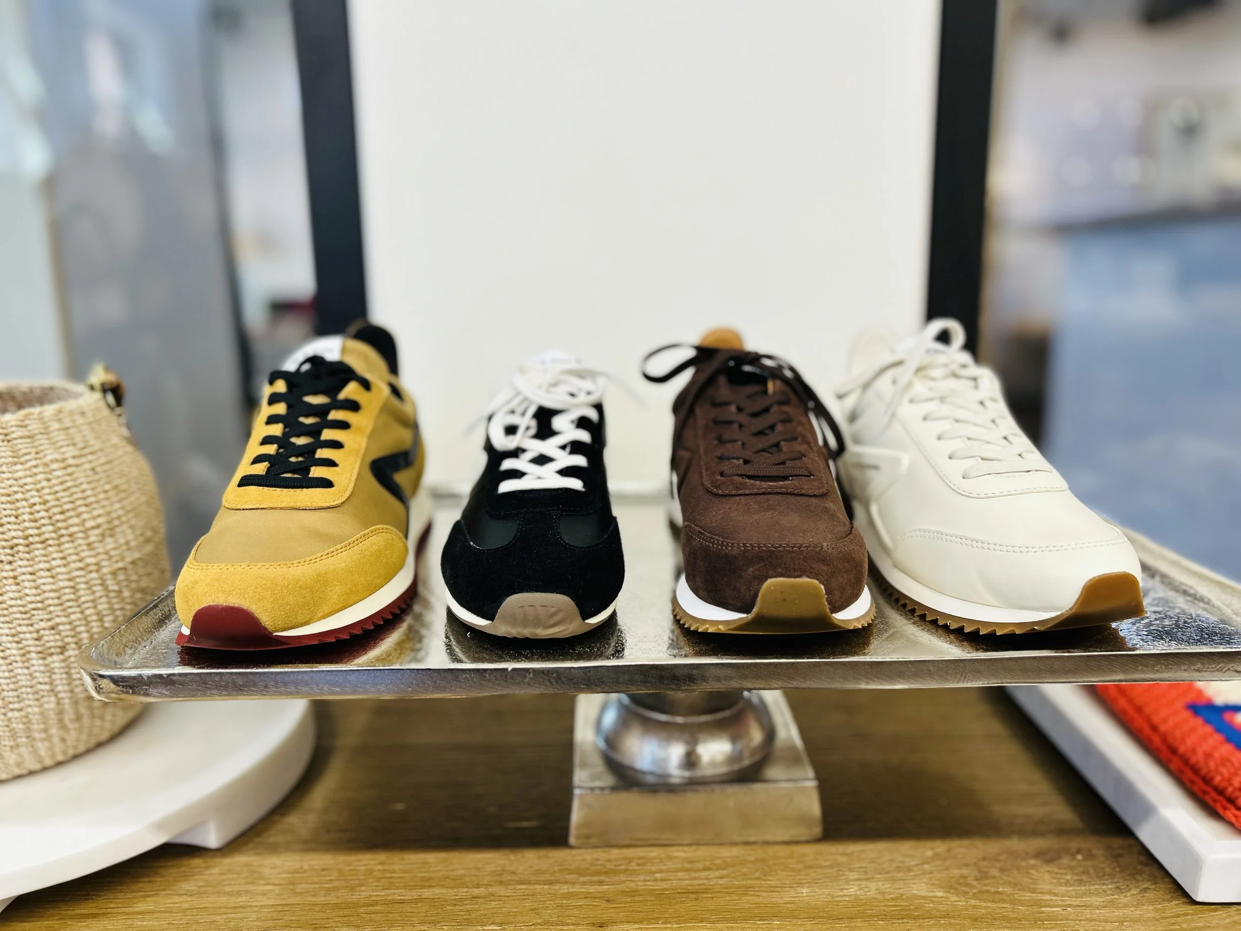 Four pairs of sneakers in yellow, black, brown, and white, displayed on a metallic stand.