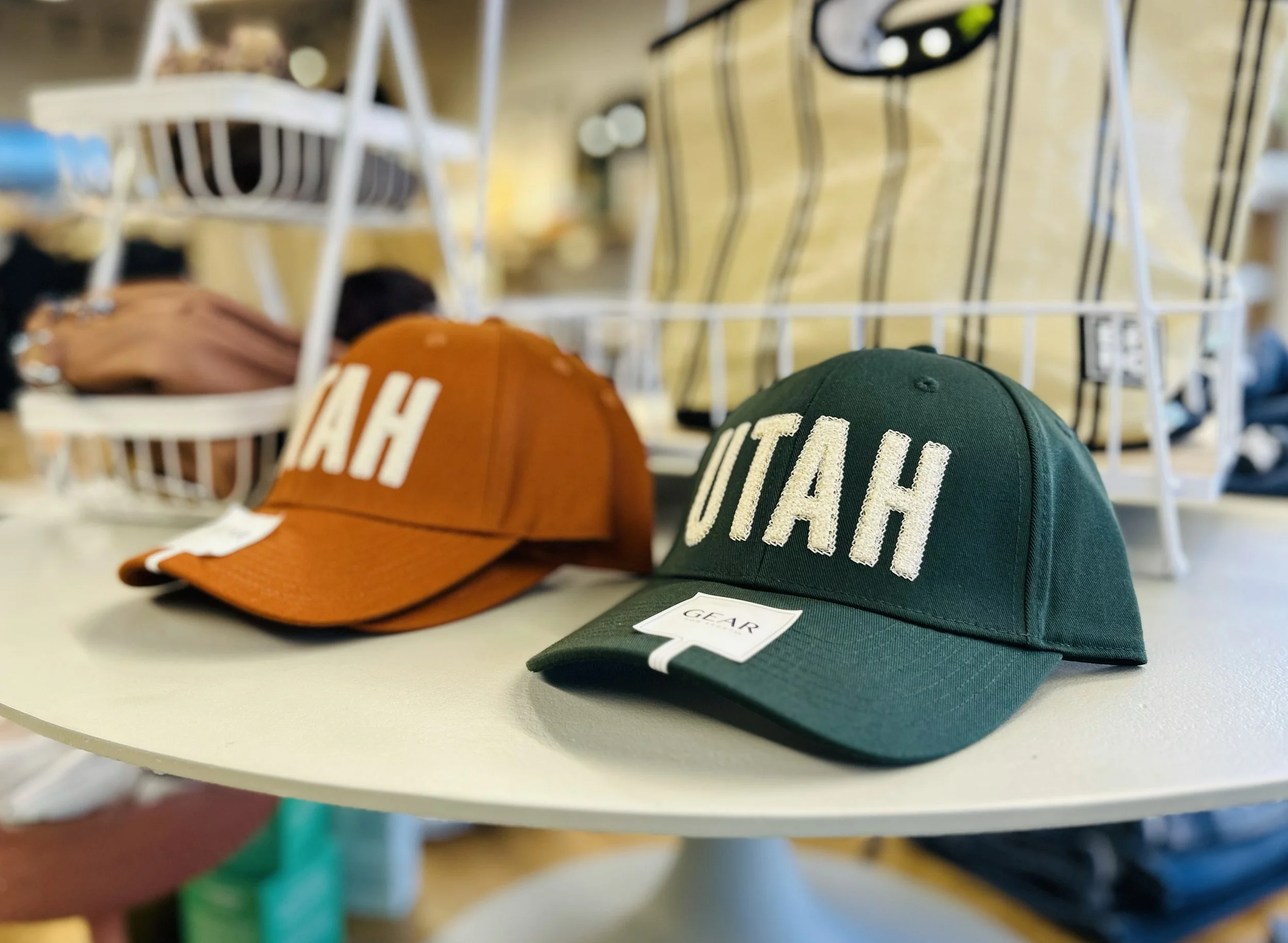 Green and orange baseball caps on a white display table, both with the word "UTAH" embroidered on the front, in a retail store setting.