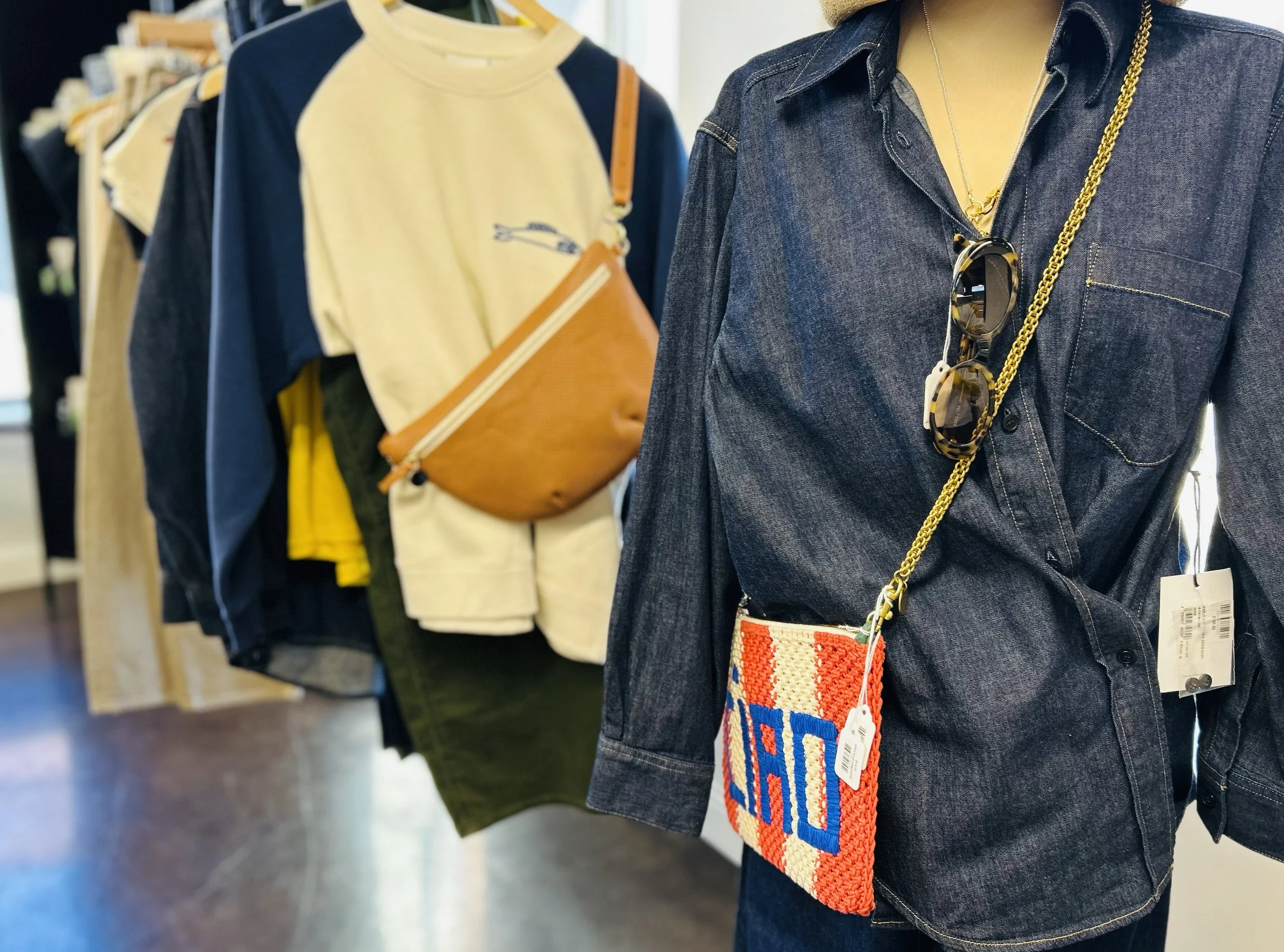 A mannequin dressed in a dark denim shirt with sunglasses hanging from a gold chain necklace. The mannequin also has a small, colorful woven bag with a shoulder strap. In the background, there are clothing racks displaying various shirts, jackets, an