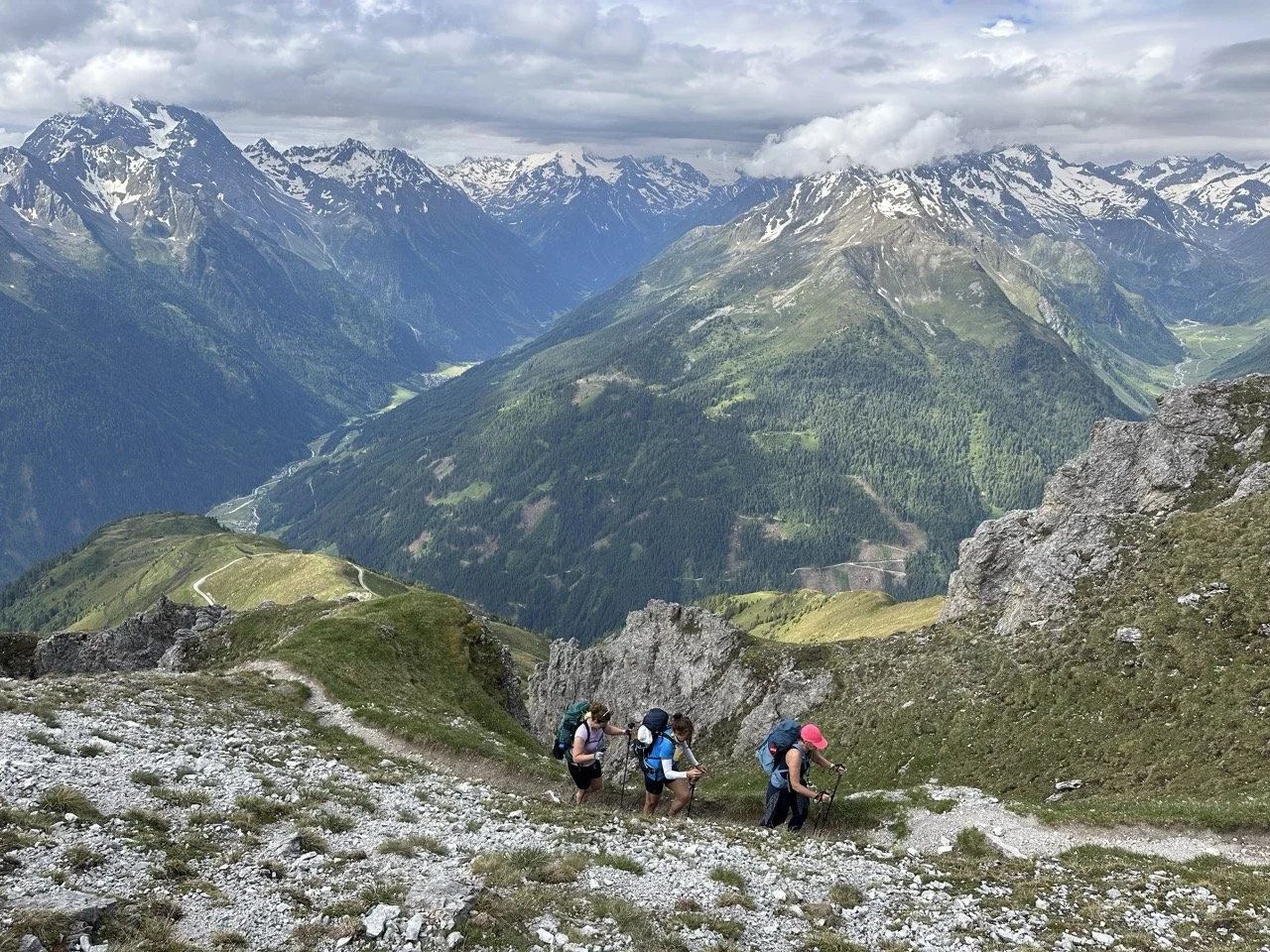 Hikers ascending a rocky mountain path with a scenic view of large mountains and a valley in the background.