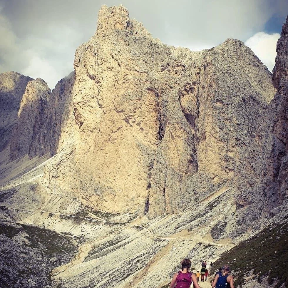 Hikers on a mountain trail, surrounded by rocky cliffs and rugged terrain under a cloudy sky.