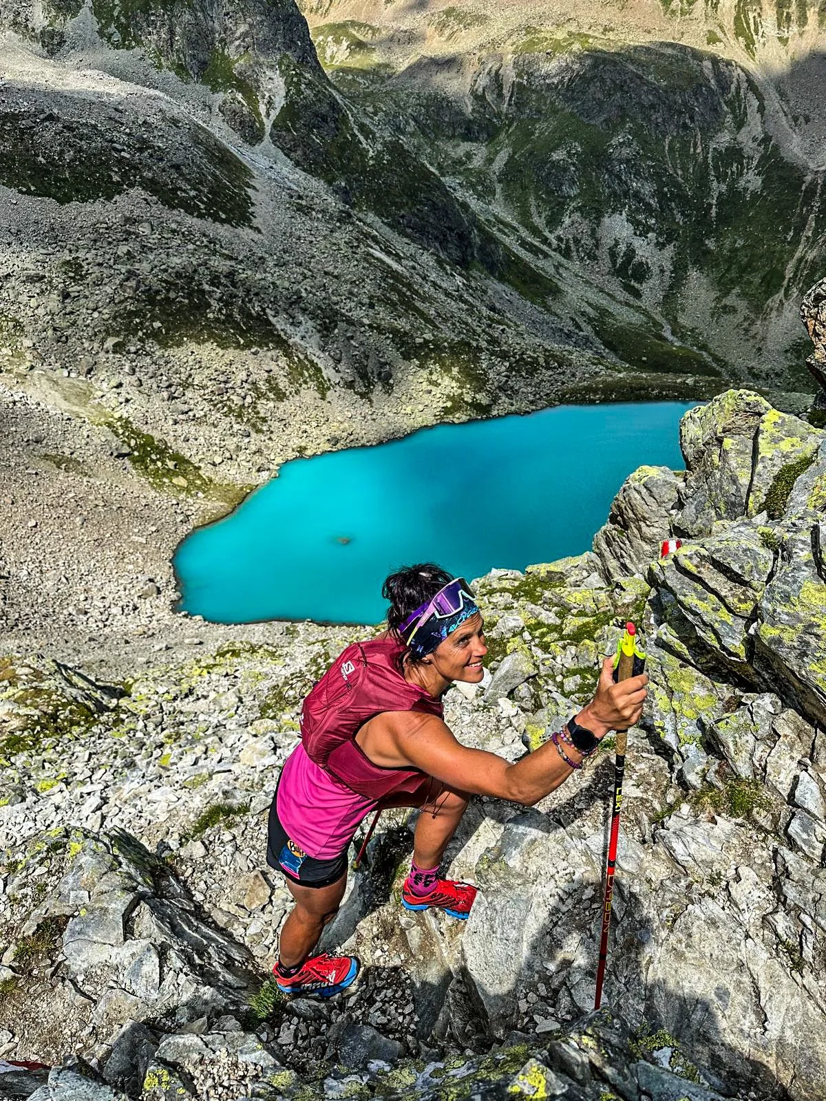 Woman climbing rocky terrain with hiking poles near a bright blue alpine lake, surrounded by mountainous landscape.