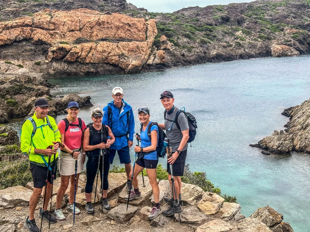 Group of hikers posing on rocky coastline with hiking poles, near clear blue water and rugged cliffs in the background.