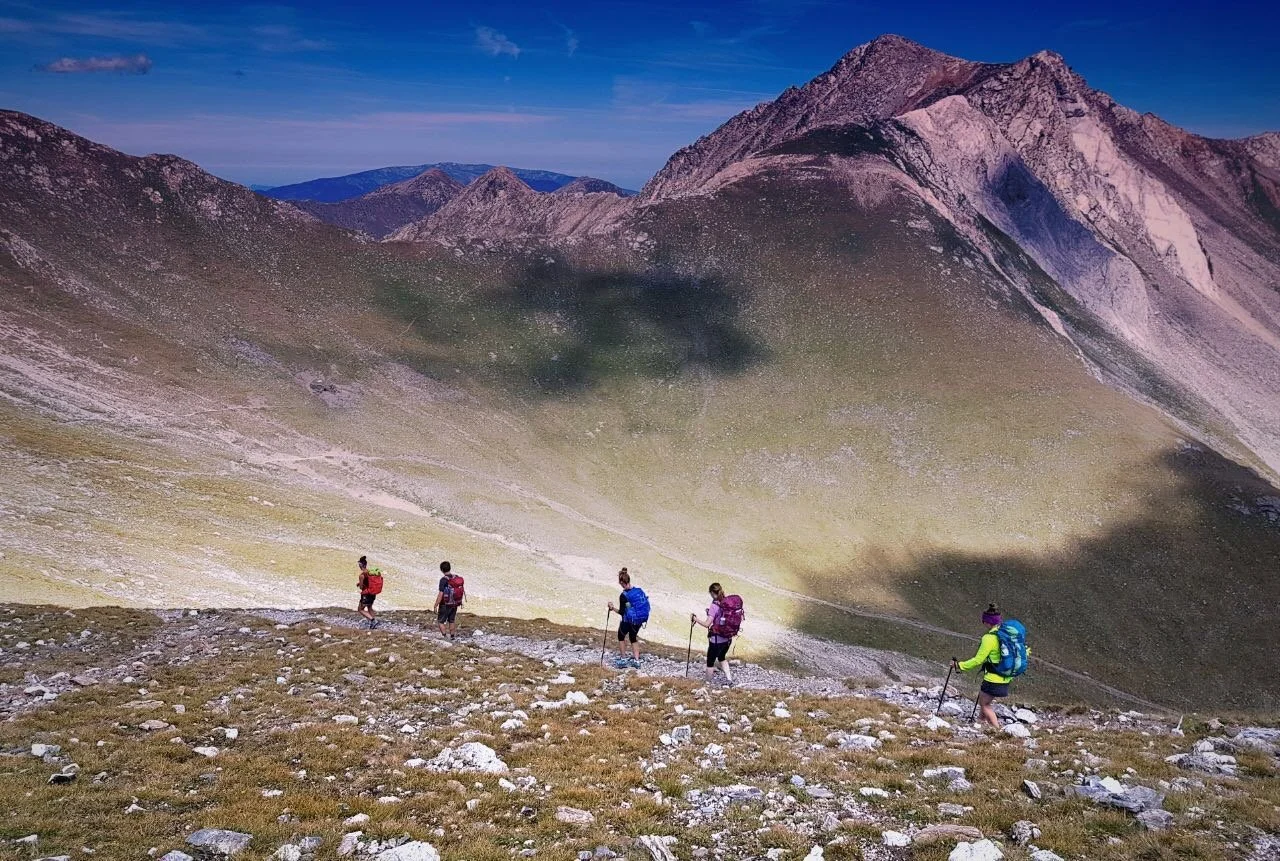 Group of hikers descending a grassy mountain slope with rocky terrain, surrounded by scenic mountain peaks under a clear blue sky.