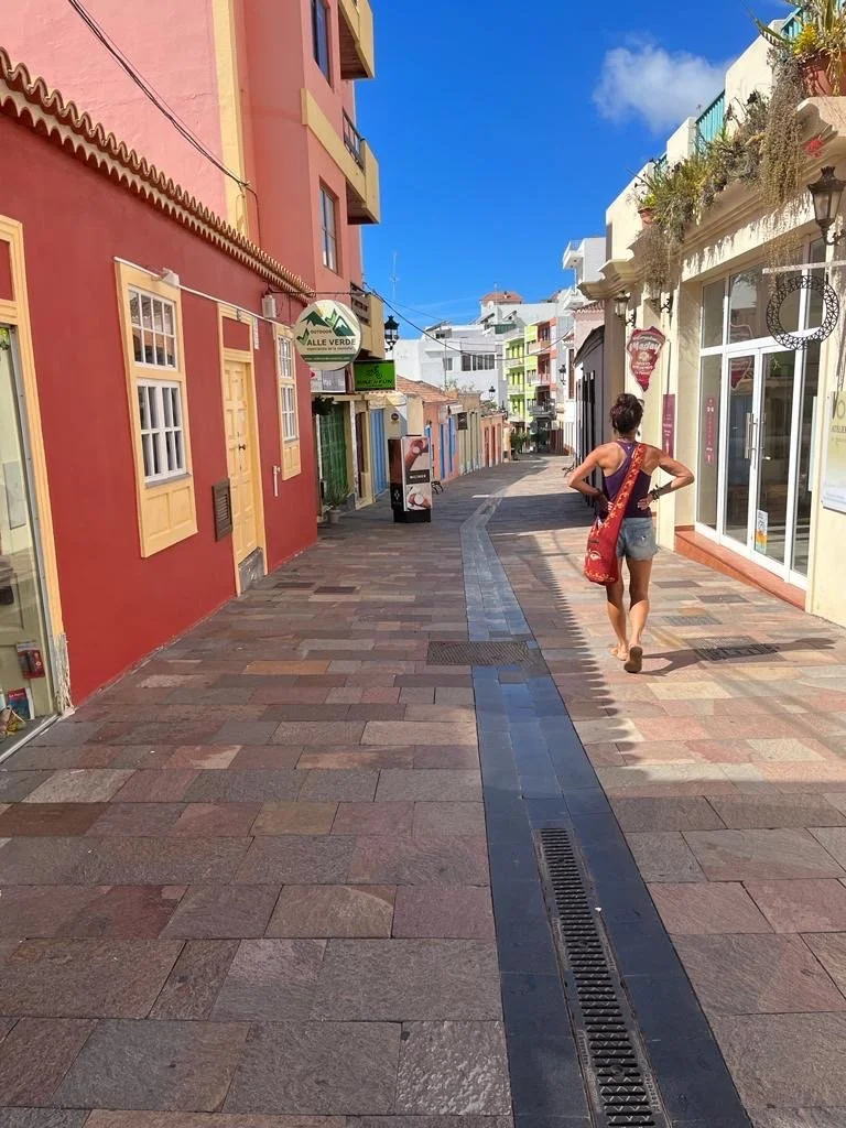 A narrow, colorful street with tiled pavement, lined with buildings painted in vibrant shades like red and yellow. A person is walking down the street carrying a red bag. The sky is clear and blue, and there is a mix of modern and traditional architecture.
