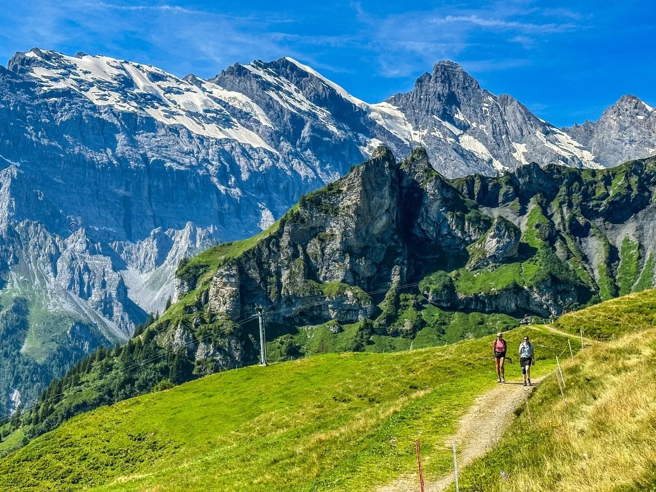 Two hikers walking on a trail in a mountainous landscape with green grass and rocky peaks.