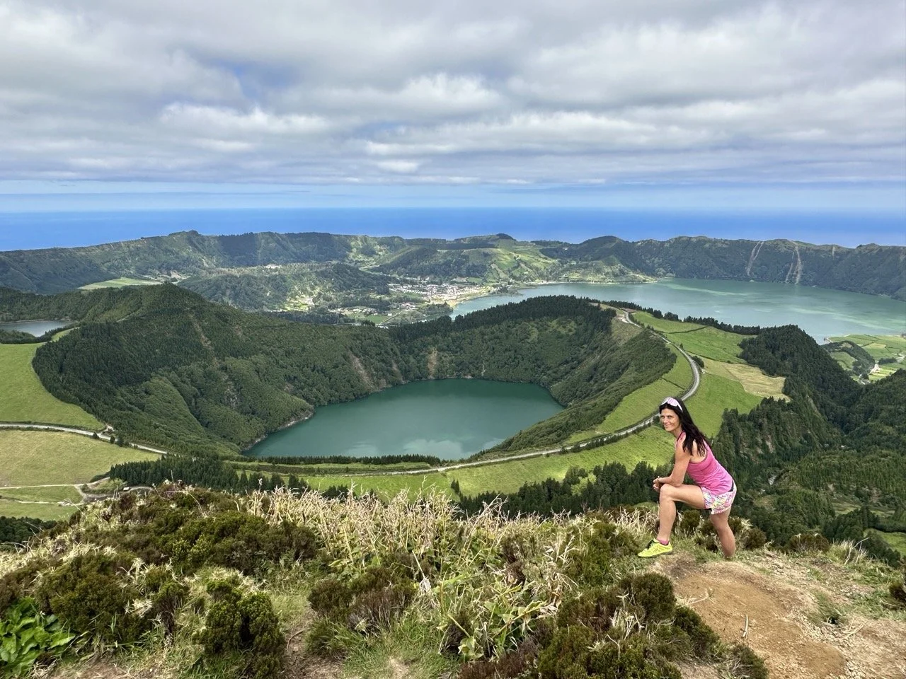 Woman posing on a hillside overlooking Sete Cidades in the Azores, with lush green landscapes and crater lakes in the background.