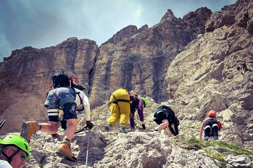 Hikers ascending a rocky mountain path with a scenic view of large mountains and a valley in the background.