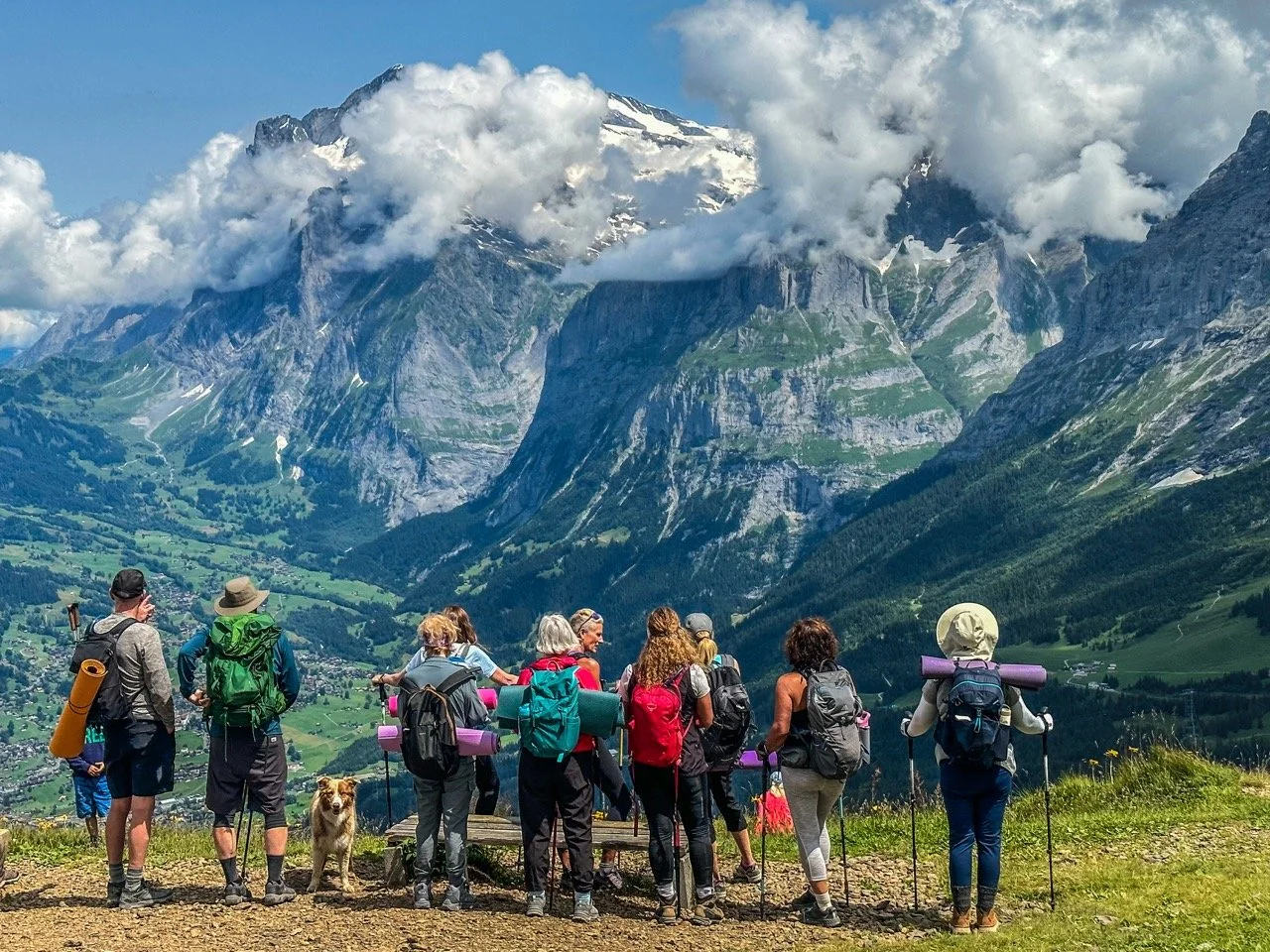 Group of hikers with backpacks and walking sticks standing on a mountain viewpoint overlooking a scenic valley and snow-capped peaks under a cloudy sky.