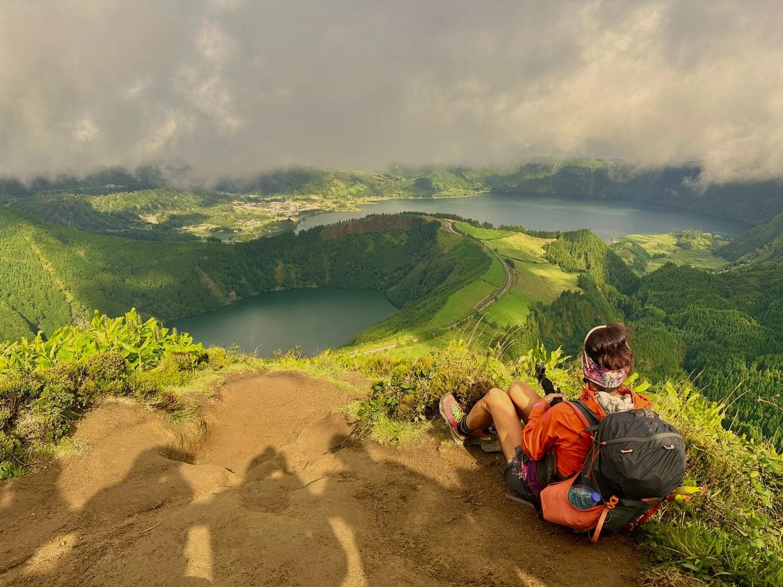 Person sitting on a hillside overlooking a verdant landscape with lakes and a cloudy sky