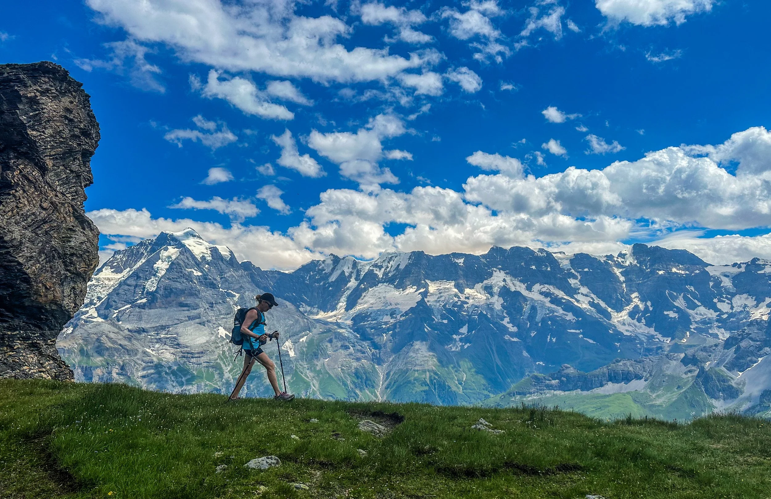 A hiker with a backpack and trekking poles walks along a grassy hillside with snow-capped mountains and a blue sky with clouds in the background.