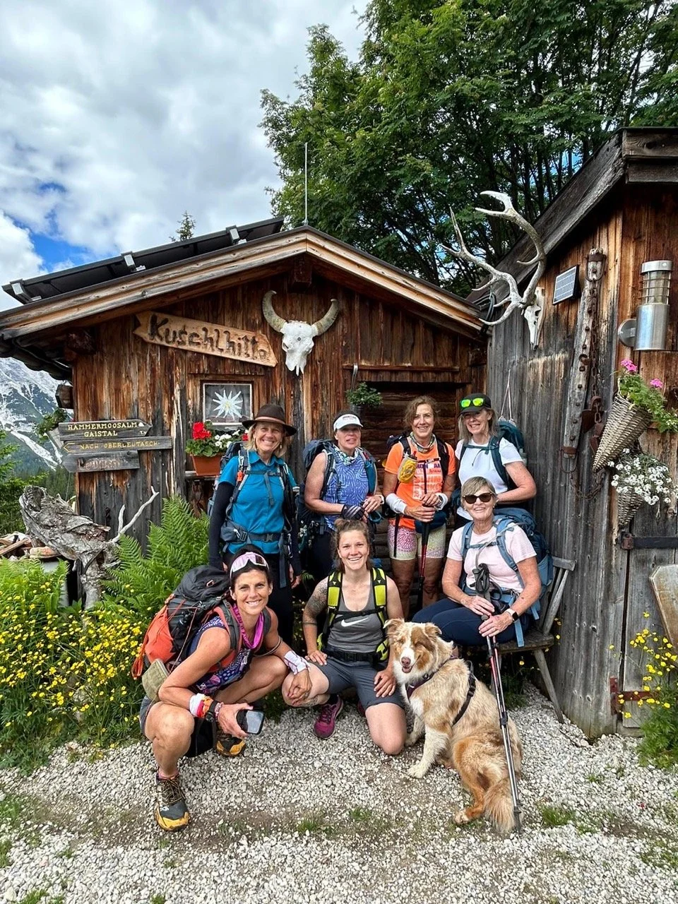 Group of hikers and a dog outside a rustic wooden cabin with mountain and tree backdrop