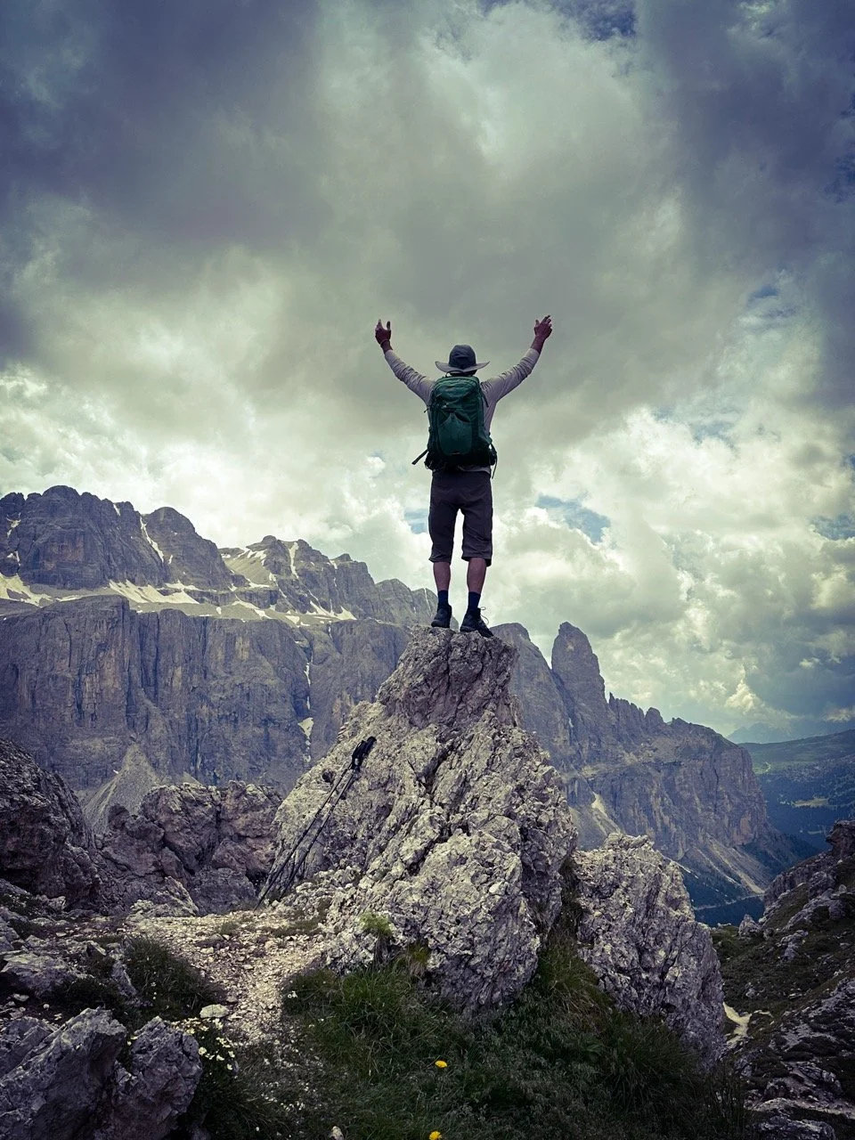 Hiker standing triumphantly on rocky peak in mountain landscape under cloudy sky.