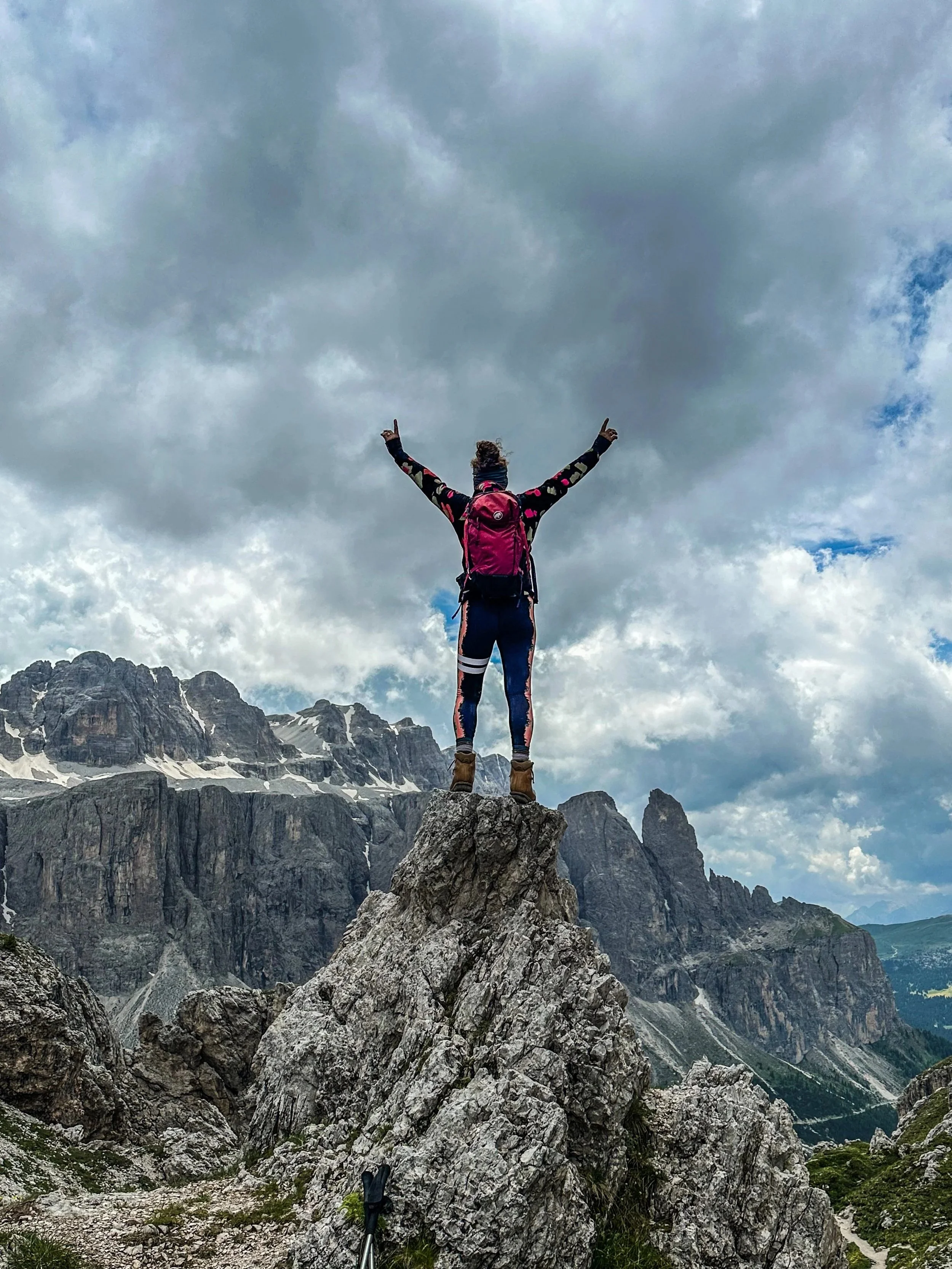 Person standing on a large rock with arms raised in victory, mountain landscape with snowy peaks and cloudy sky in the background.