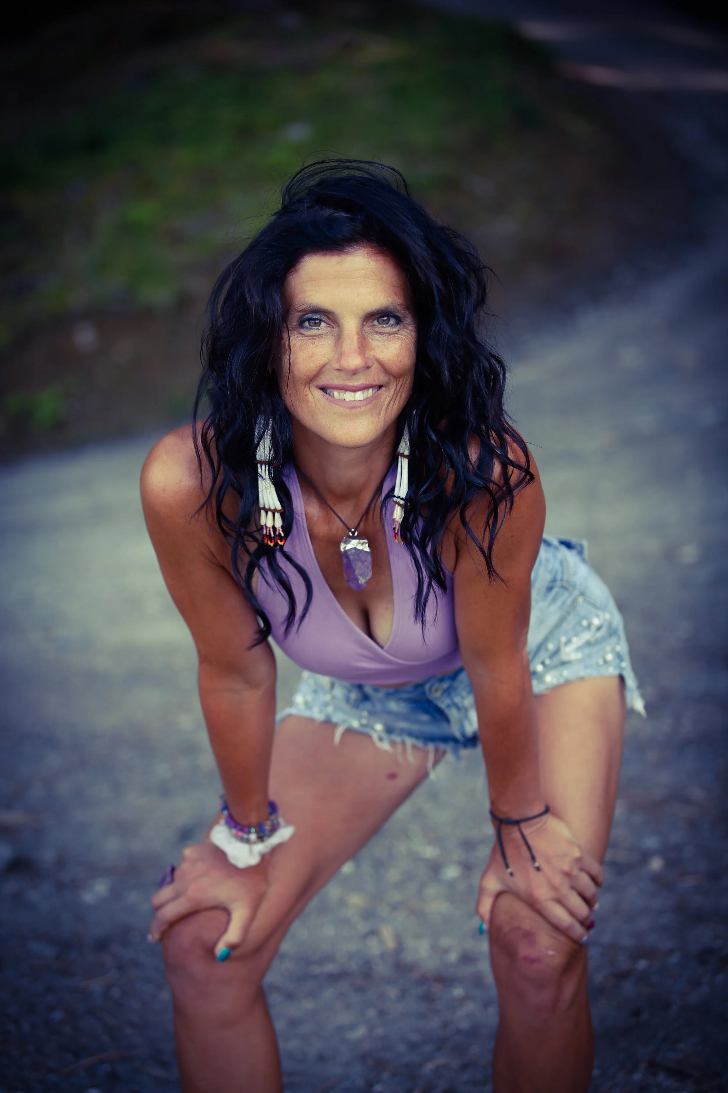 Woman with dark hair, wearing a lavender tank top and denim shorts, leaning slightly forward with hands on knees, smiling outdoors.