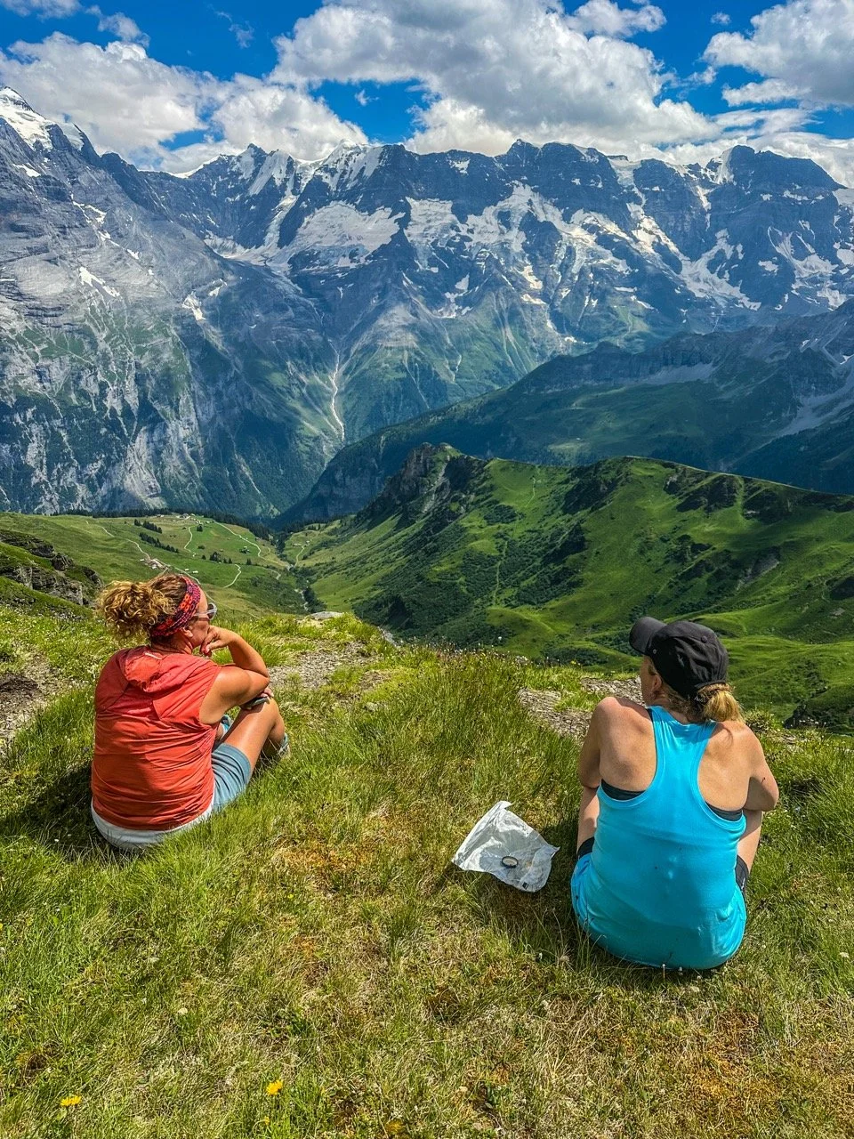 Two hikers sitting on a grassy hilltop with a panoramic view of snow-capped mountains and a clear blue sky.