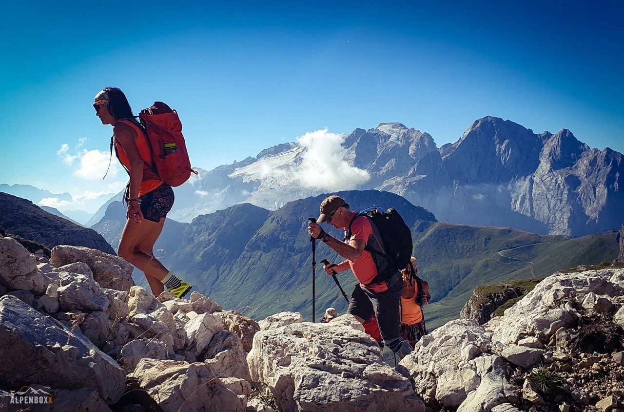 Hikers climbing rocky terrain with mountain backdrop, clear blue sky.