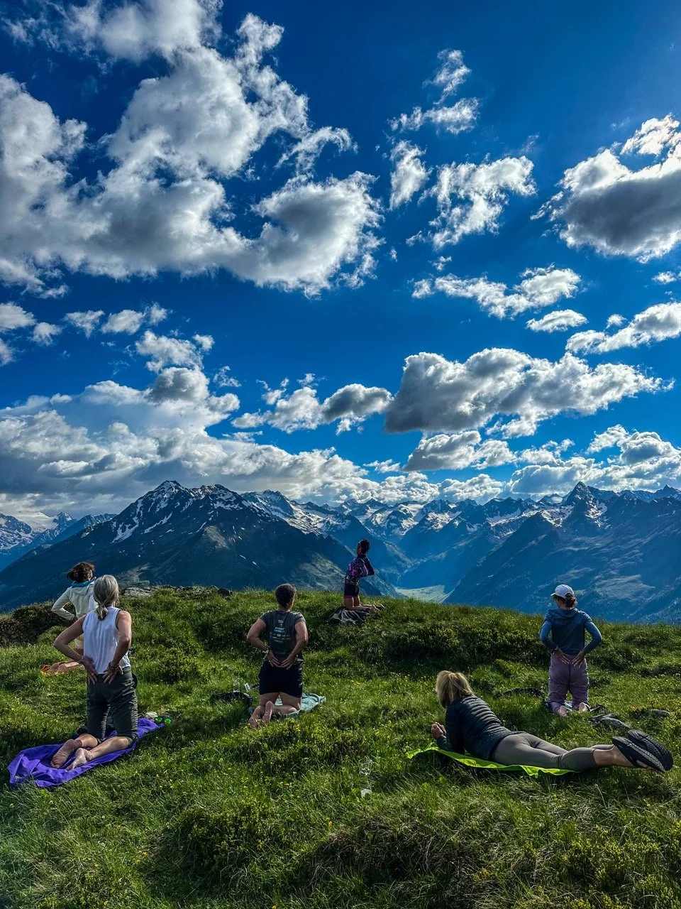 Group of people in yoga poses on a grassy hill with snowy mountains in the background under a partly cloudy sky.