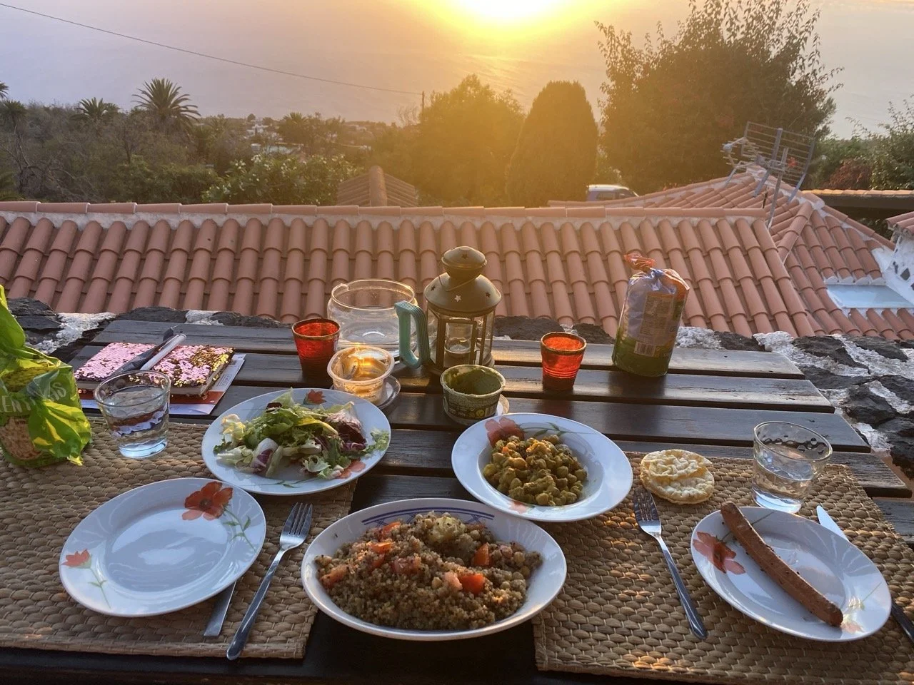 Outdoor dining setup with a variety of dishes on a table, including salads, grains, sausage, and condiments, with a scenic sunset view and rooftops in the background.