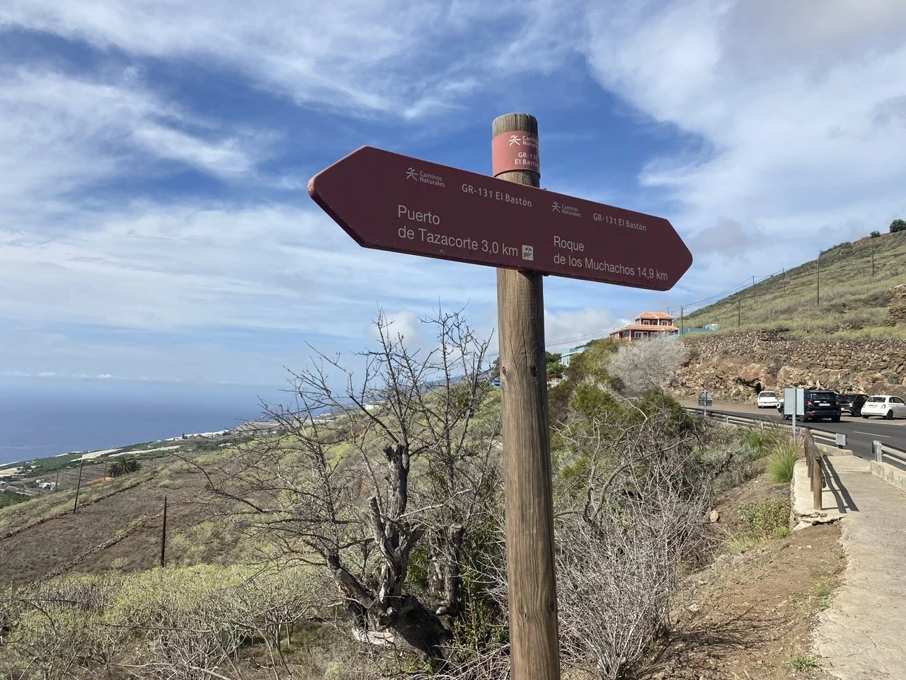Signpost on a scenic road indicating directions to Puerto de Tazacorte and Roque de los Muchachos in a hilly landscape.