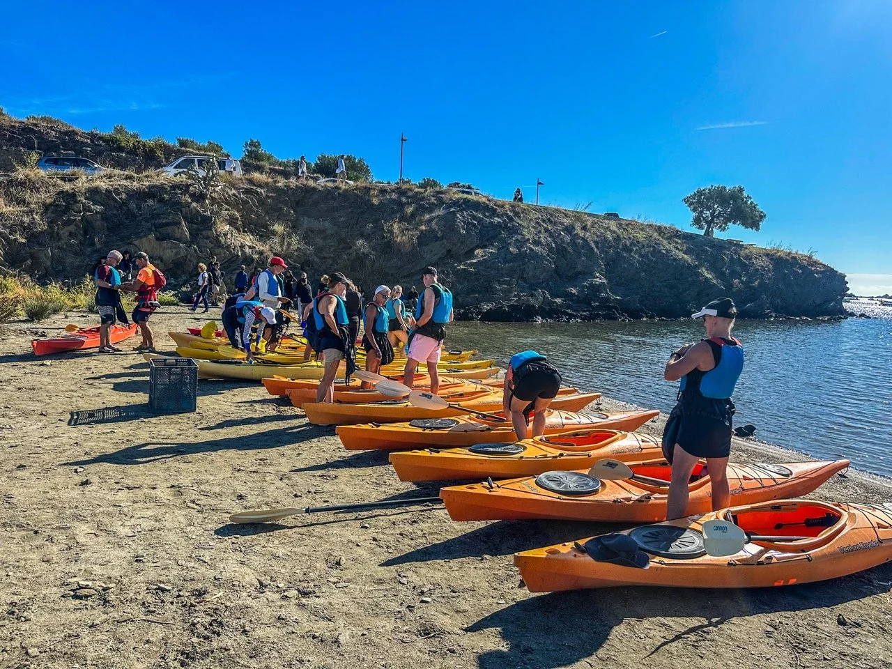 A group of people preparing kayaks on a sandy shore near a water body under a clear blue sky. They are wearing life jackets and organizing equipment, with a rocky hill in the background.