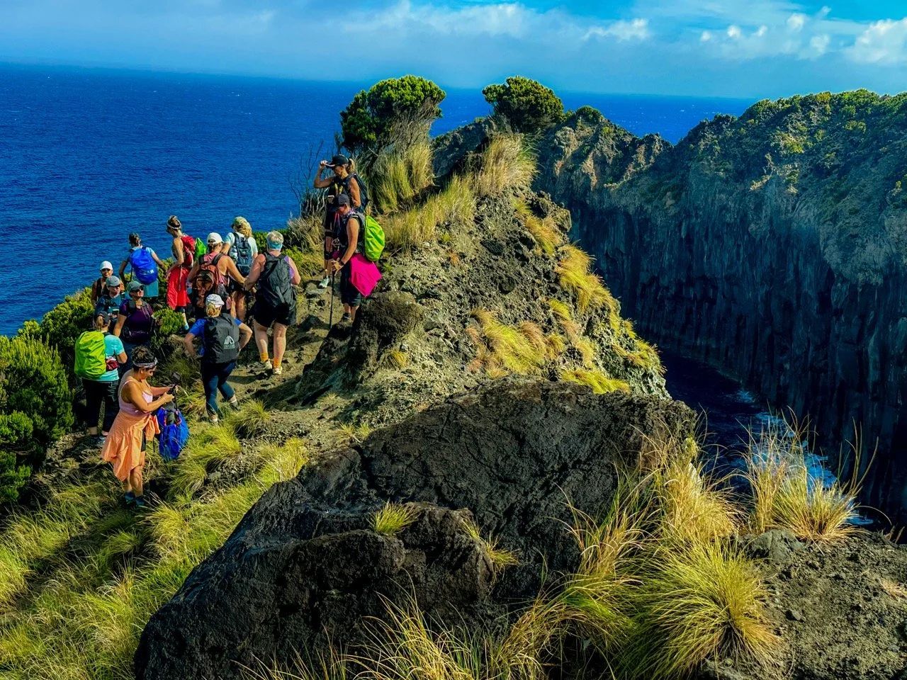 A group of people hiking on a rocky cliff with ocean views in a scenic coastal area under a partly cloudy sky.