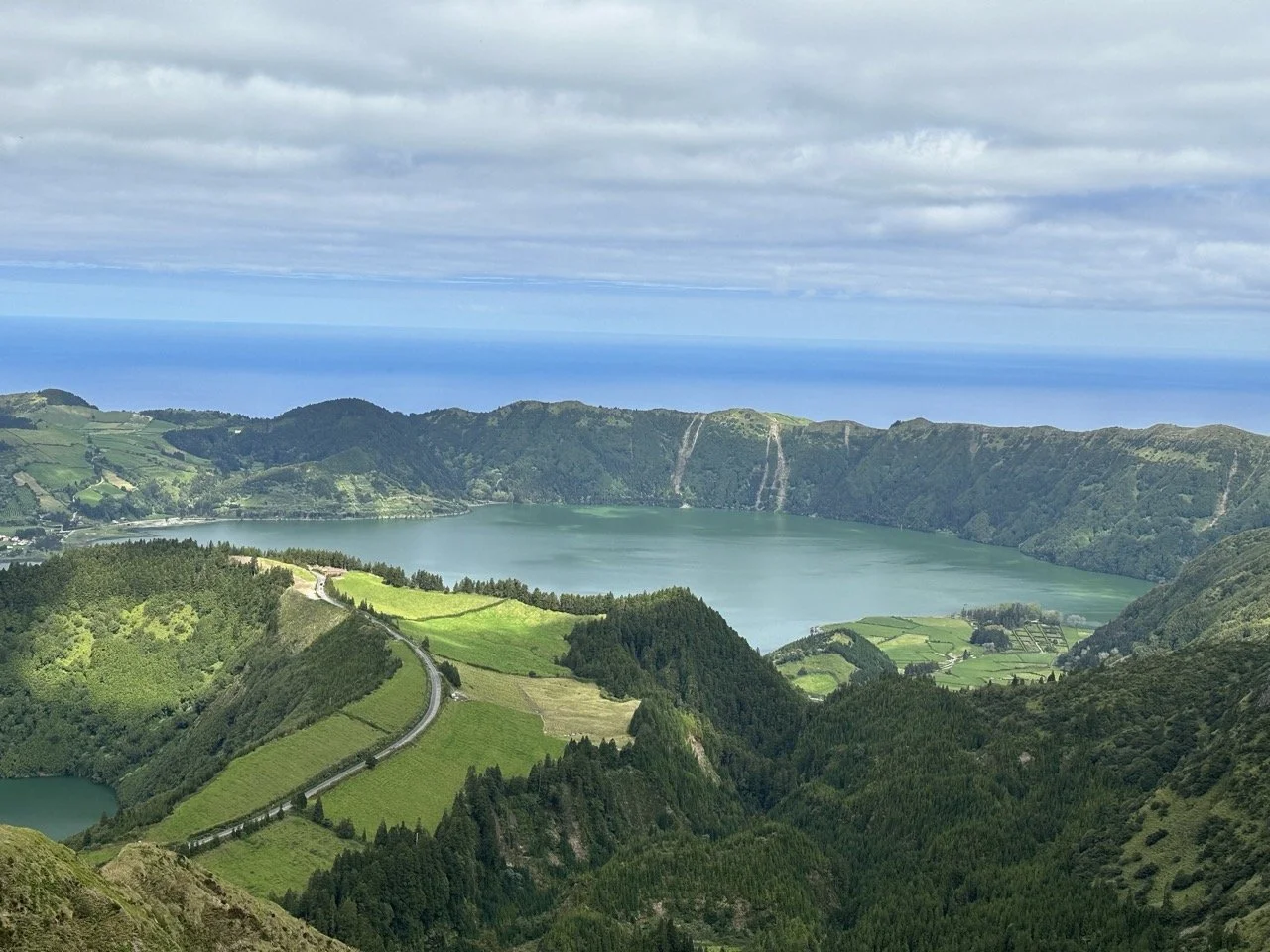 Panoramic view of greenery, hills, and a lake under a cloudy sky, with the ocean in the background.