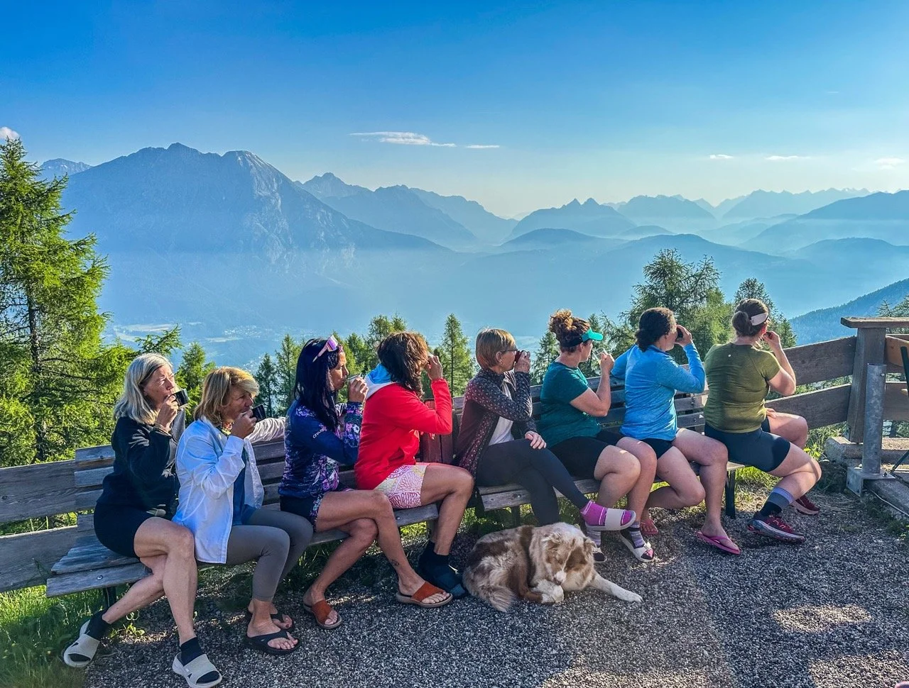 Group of people sitting on a bench with a scenic mountain view, drinking from mugs, and a dog lying on the ground.