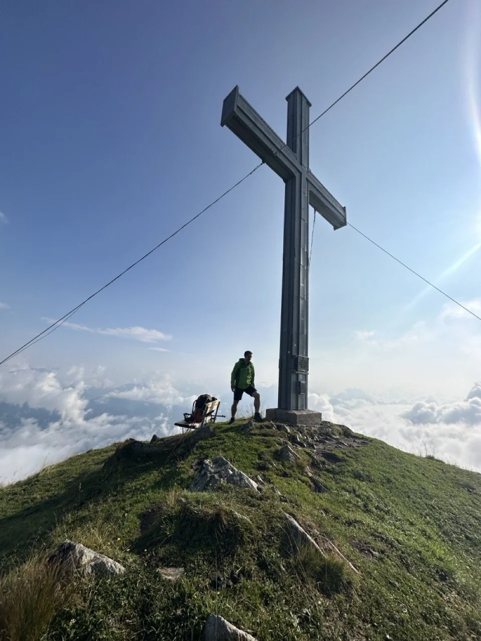 Person standing near a large cross on a mountain peak, surrounded by clouds and clear blue sky.