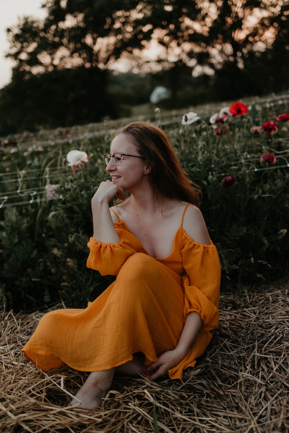 A woman in a yellow dress sits in a field of flowers, smiling and looking to the side, with trees in the background at sunset.
