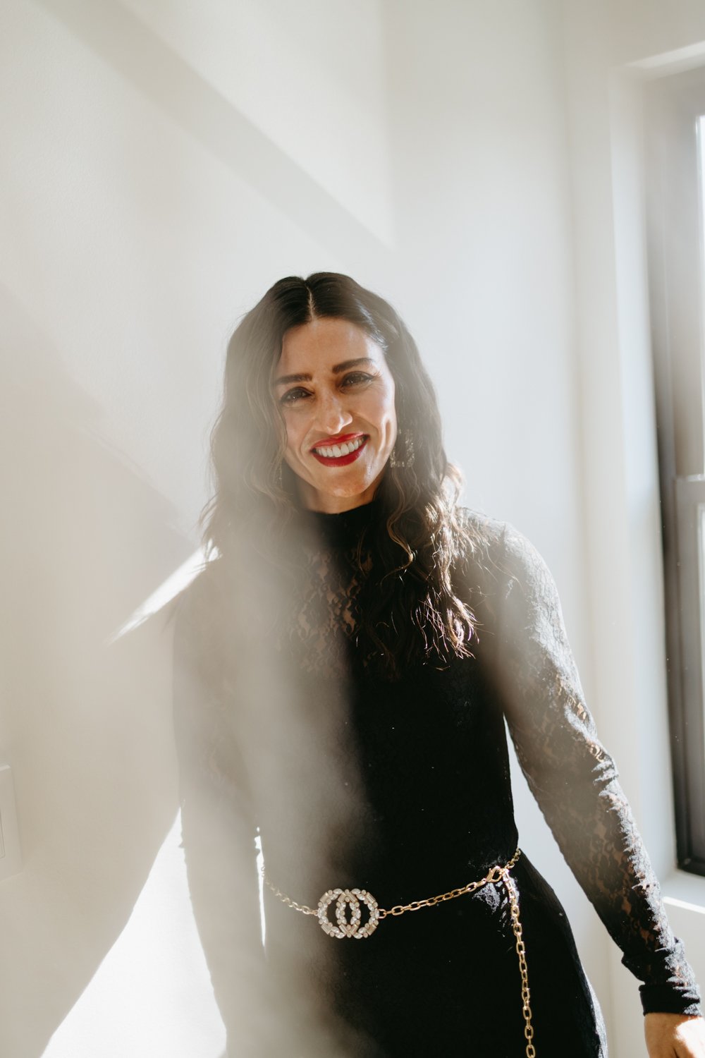 Woman in black lace dress with chain belt, smiling in sunlight.