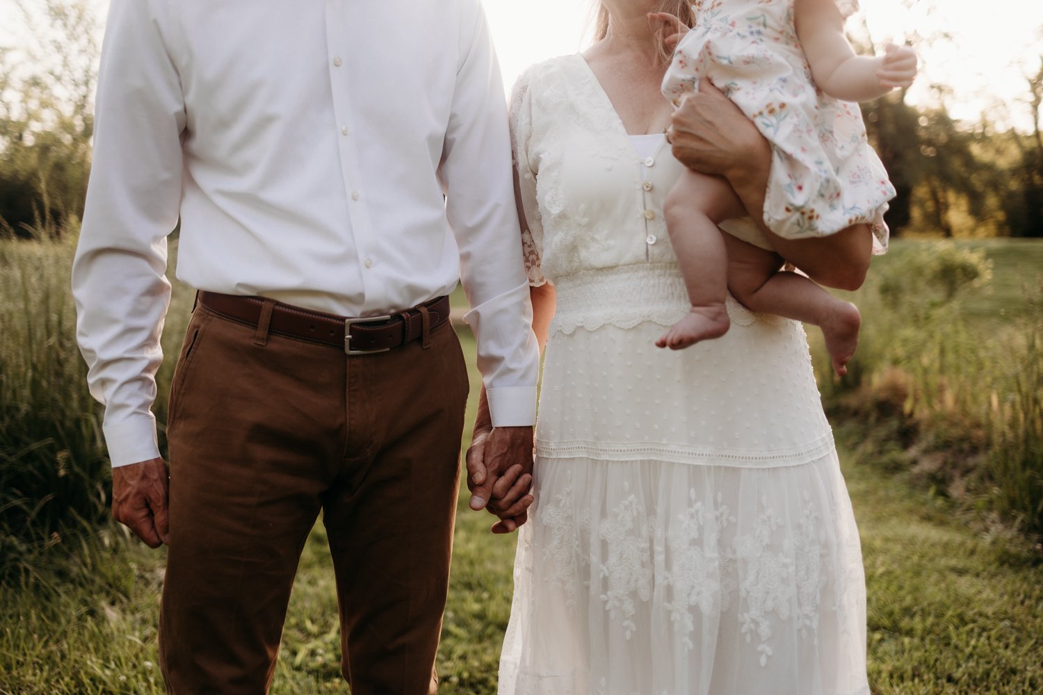 A man and woman holding hands, with the woman carrying a baby, standing on grass outdoors. The man wears a white shirt and brown pants, while the woman wears a white dress.
