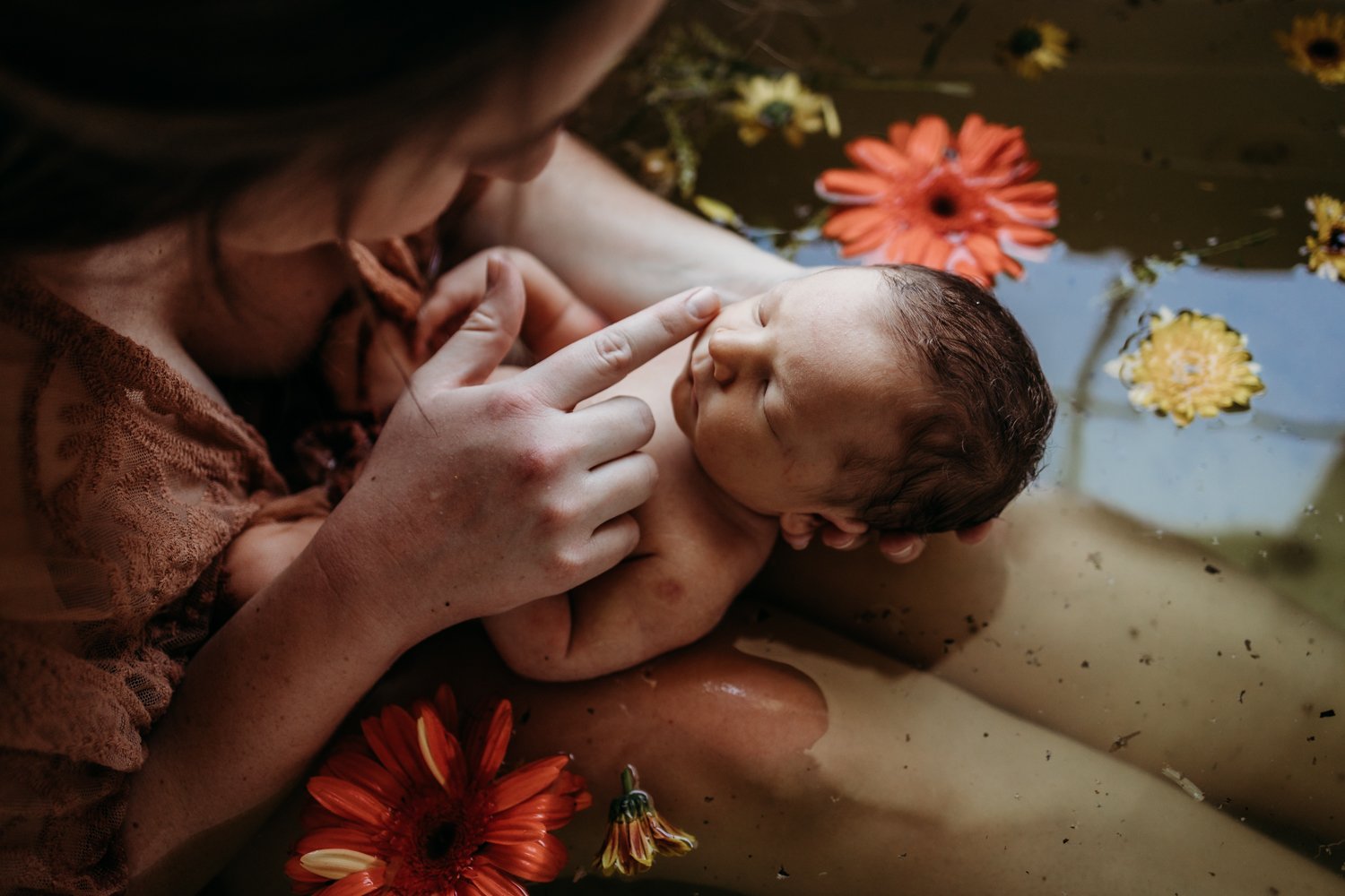 Mother holding newborn baby in water with floating flowers.
