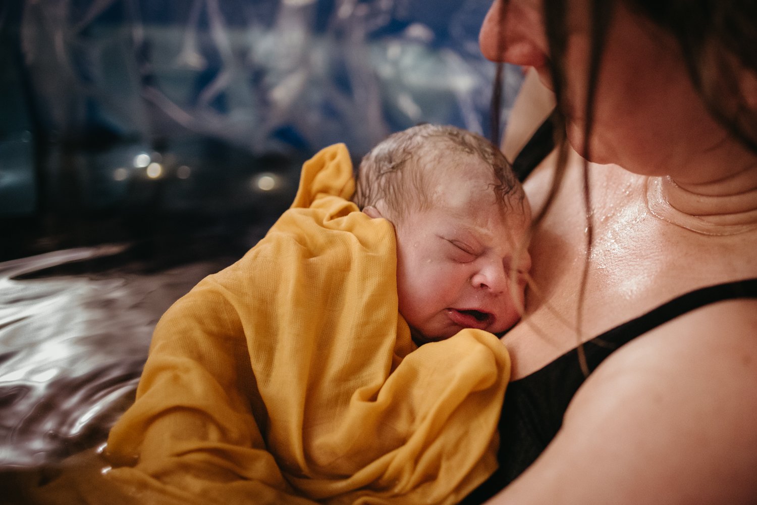 A newborn baby wrapped in a yellow blanket, cradled against the chest of an adult, likely the mother, in a watery environment, possibly during a water birth.