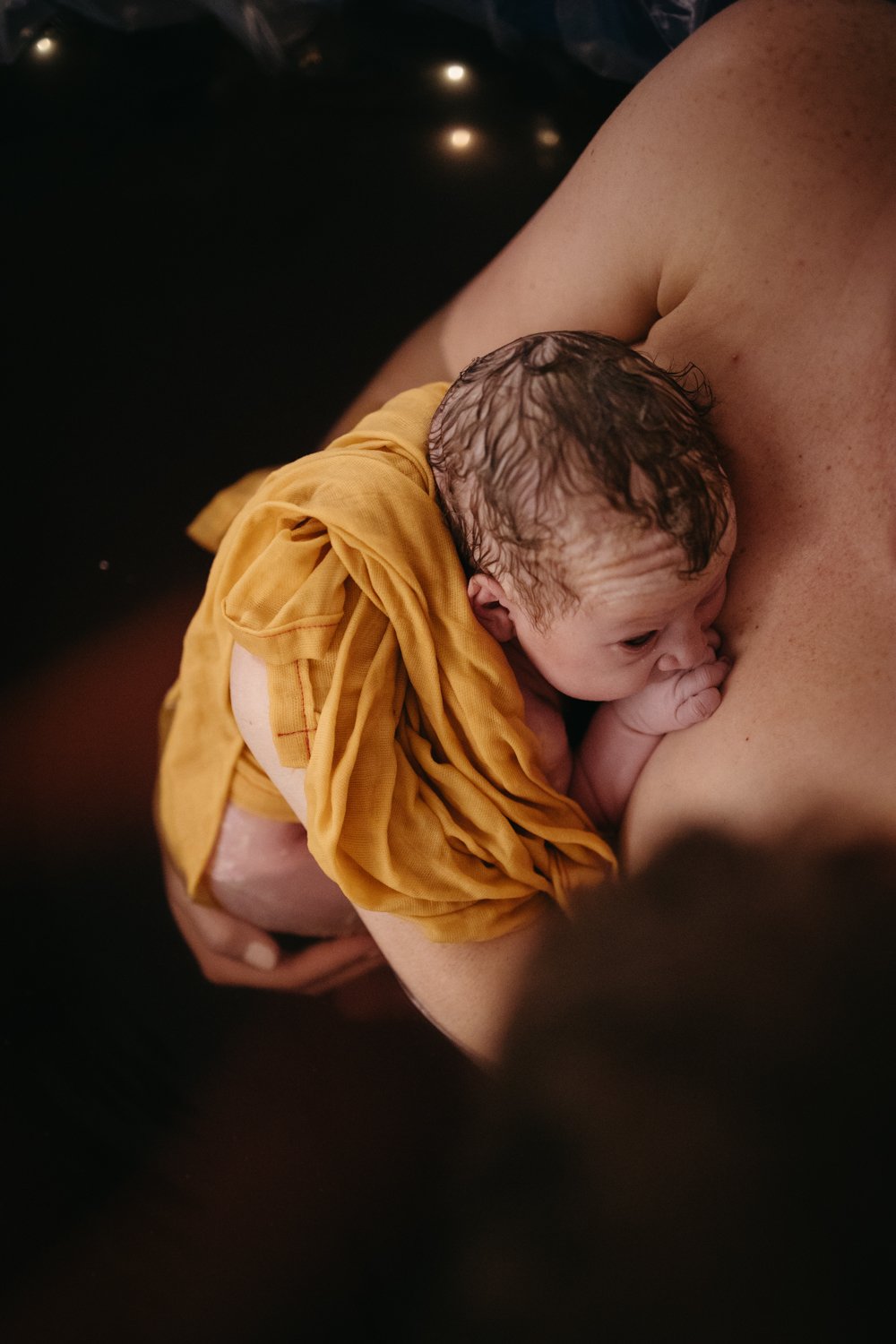 Newborn baby wrapped in a yellow cloth held by a mother in a birthing pool.