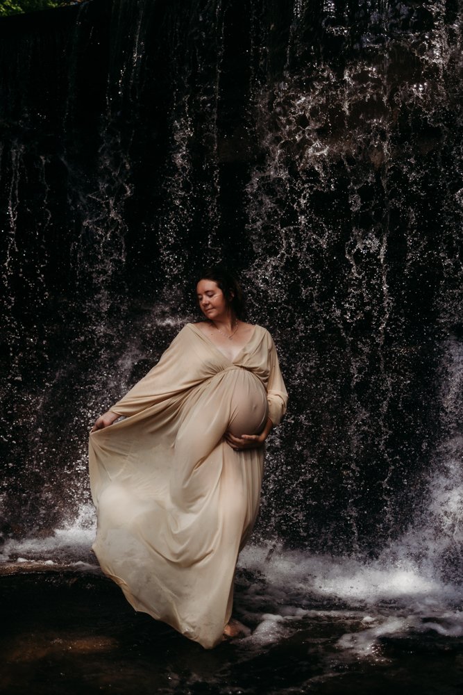 A pregnant woman in a flowing beige dress standing in front of a waterfall, holding her belly and looking serene.