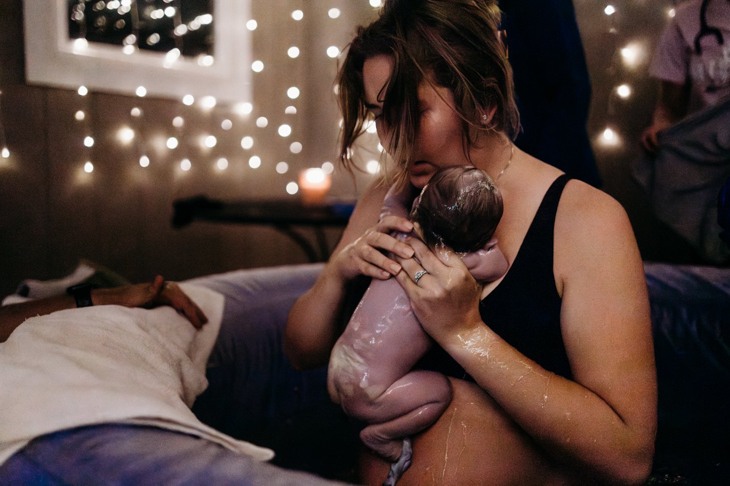Woman holding newborn after water birth in a dimly lit room with string lights.