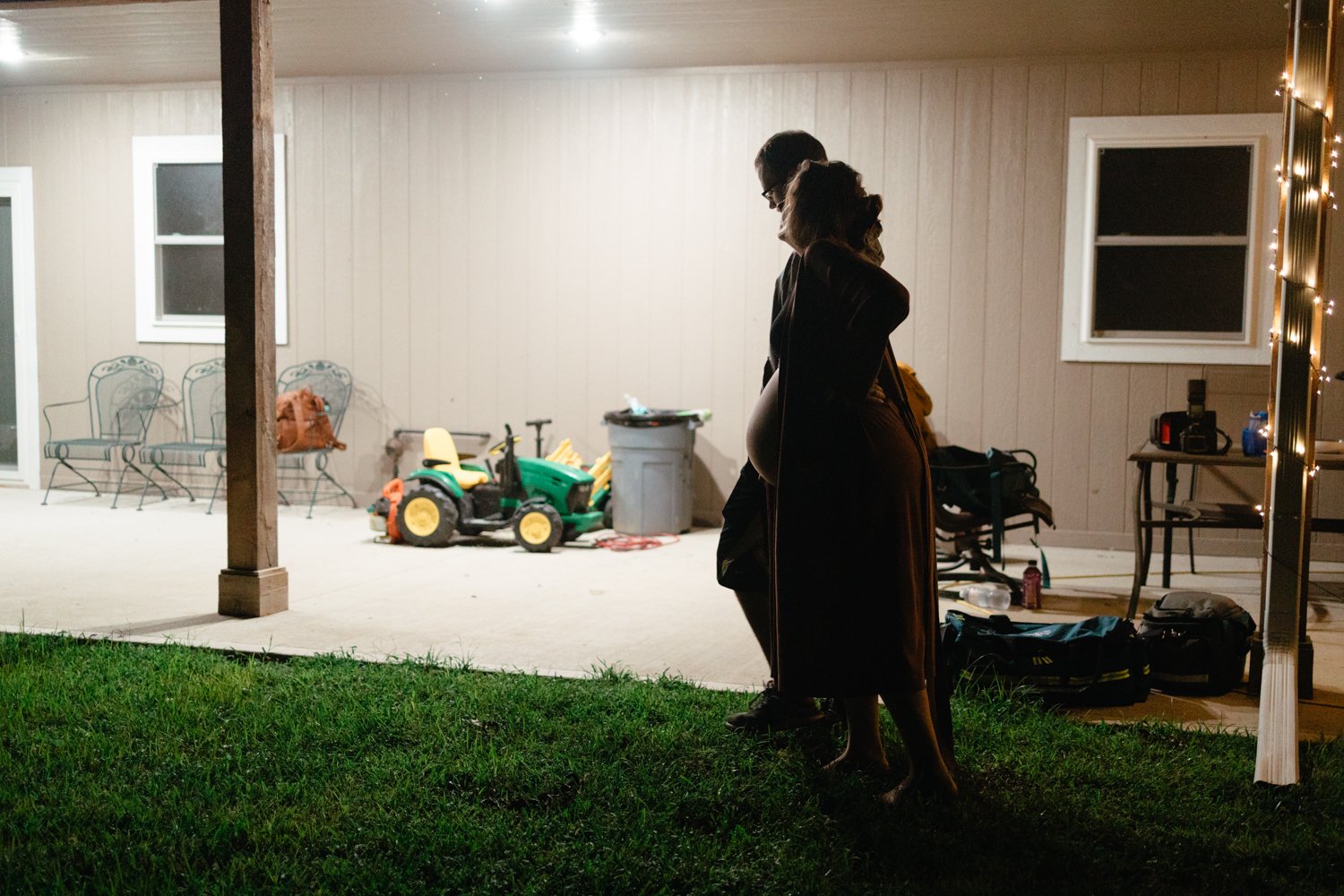 A dimly lit porch with a pregnant woman standing beside another person at night. The porch has a green toy tractor, patio chairs, bags, and string lights on a post.