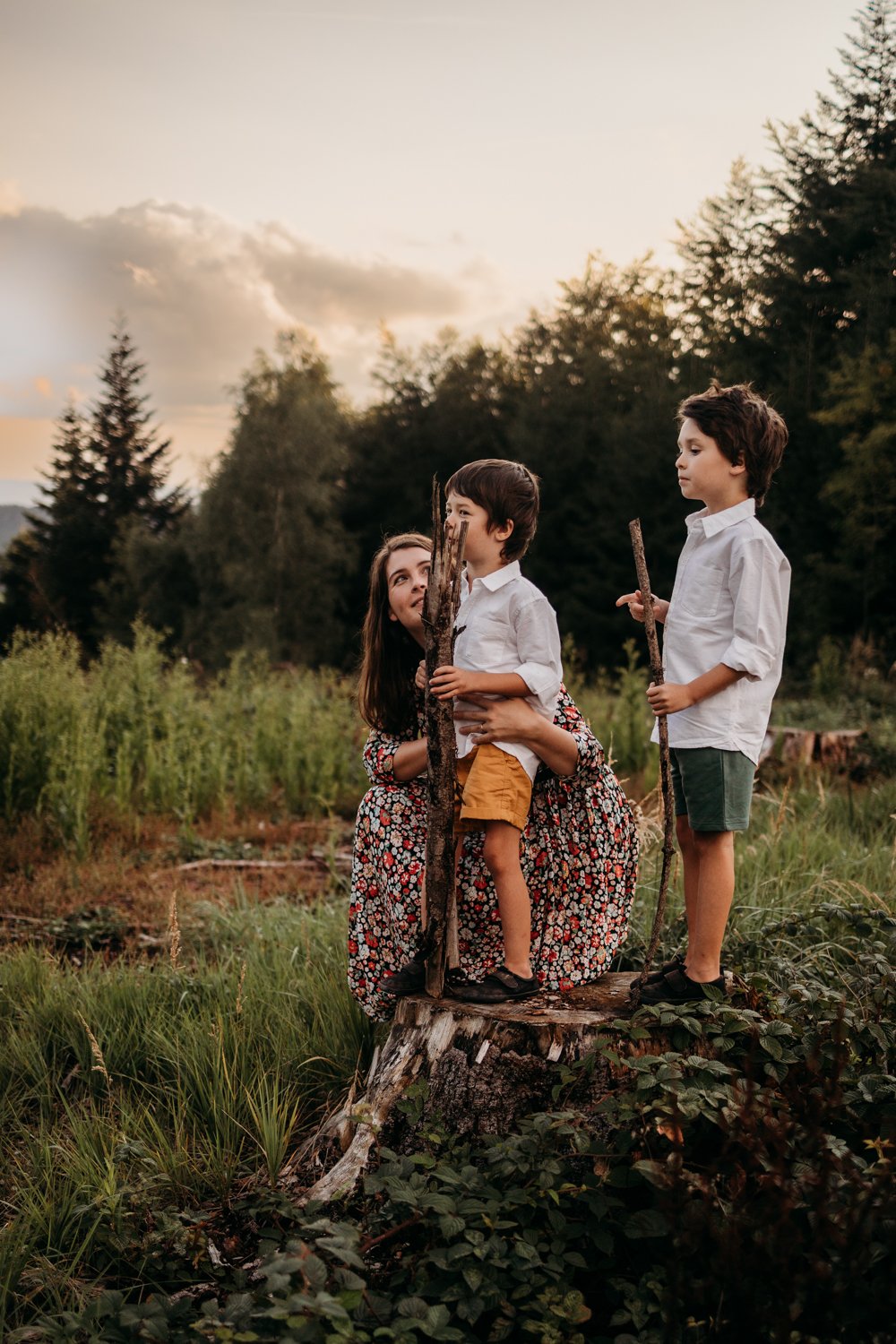 A woman and two children playing in a forest clearing, holding sticks, wearing casual summer clothes.