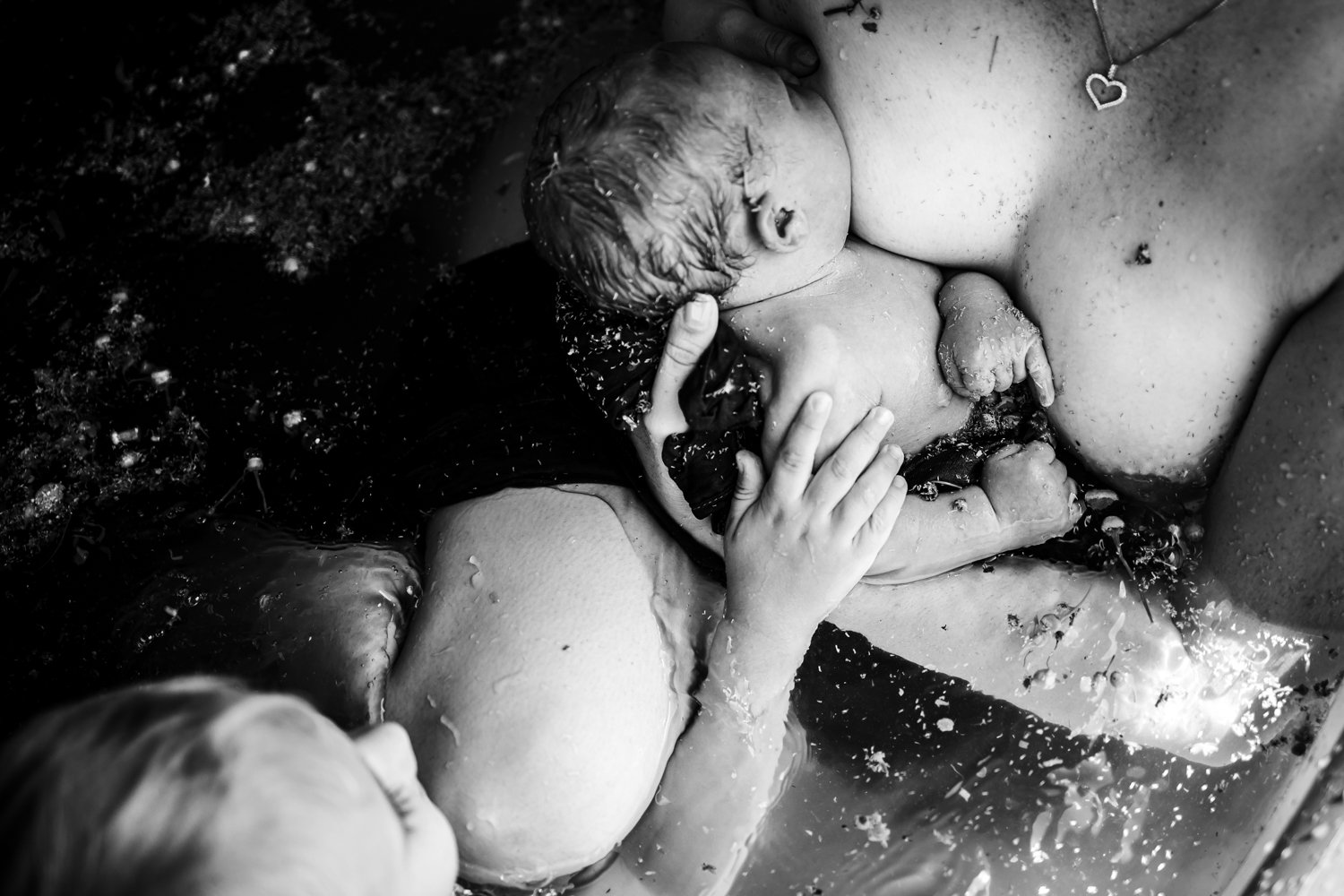 Black and white image of a newborn baby during a water birth, held close to a woman's chest, with foliage and water visible.