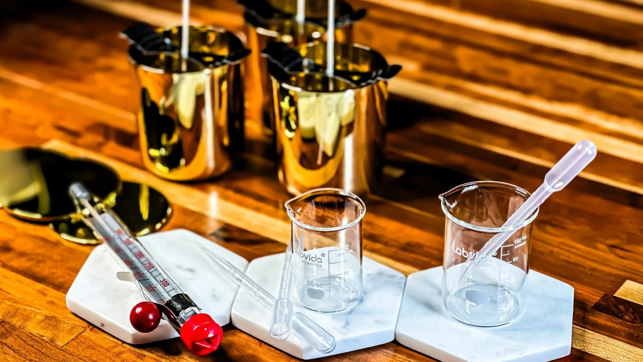 Laboratory glassware including beakers, a pipette, and a thermometer on a white marble slab on a wooden table, with gold candle vessels in the background.