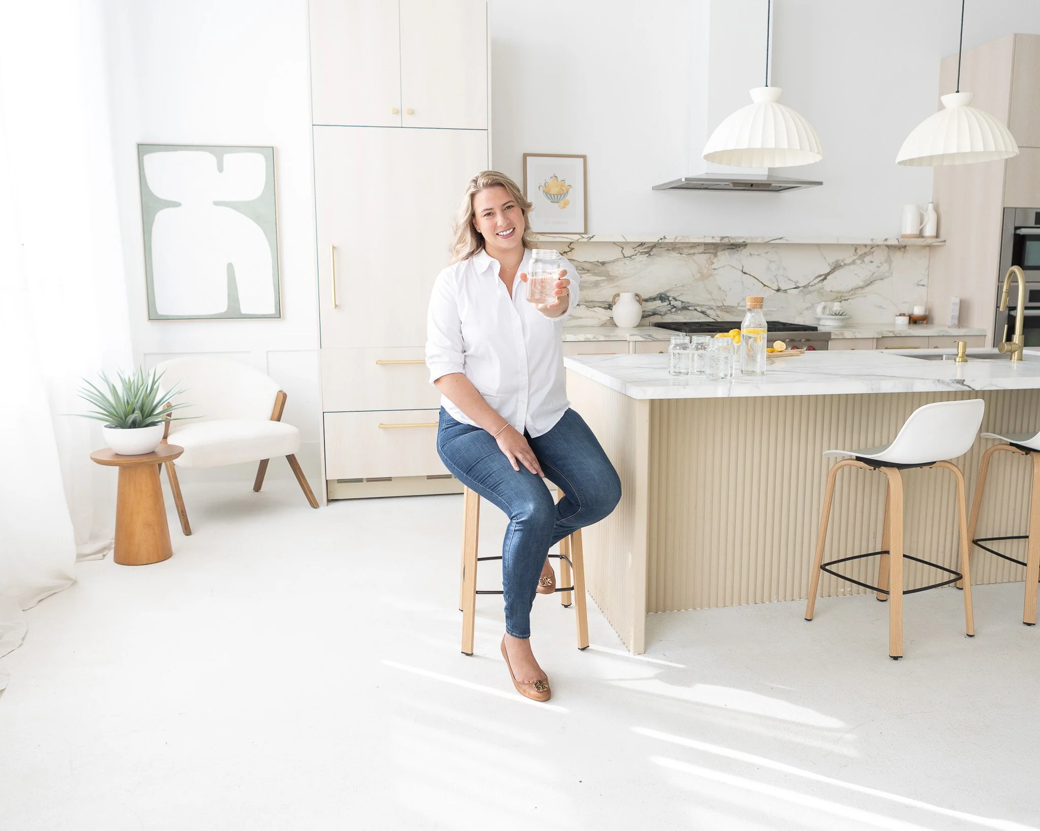 Woman sitting on a stool in a modern, airy kitchen holding up a glass of water and smiling.