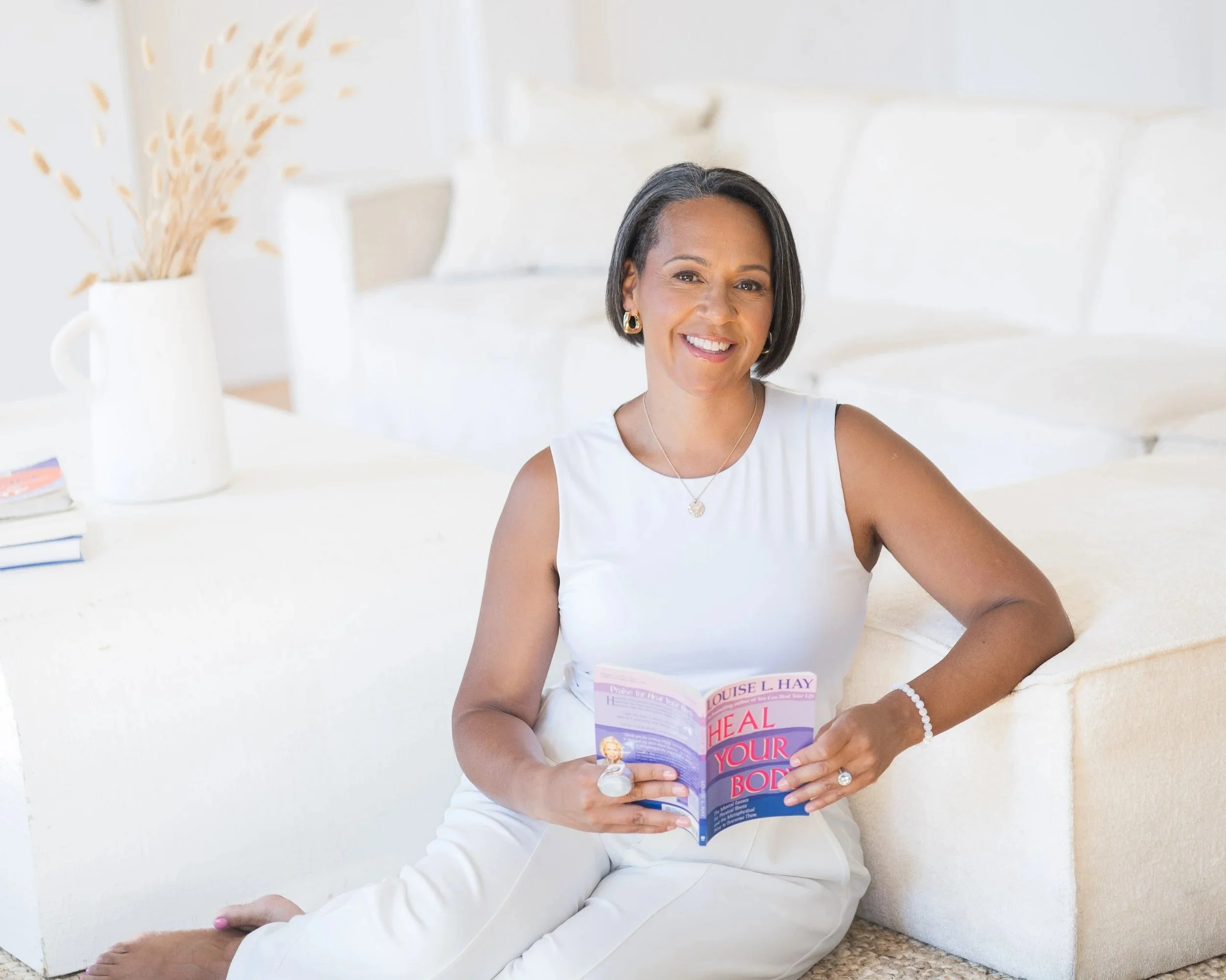 Smiling woman sitting on the floor reading a book titled 'Heal Your Body' in a bright, minimalist room with beige furniture and decorative vase.