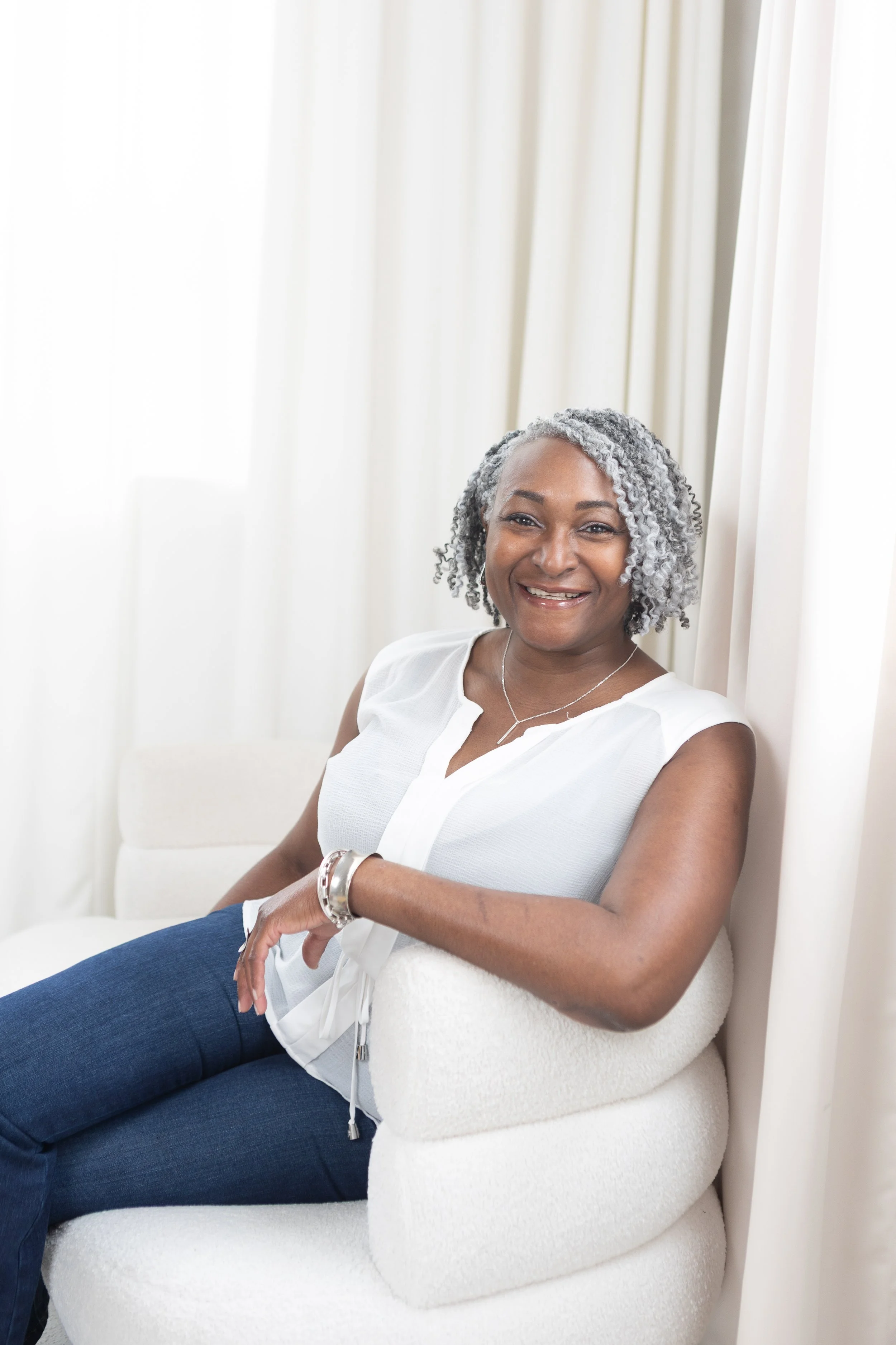 Smiling woman with gray curly hair sitting on a white chair by cream-colored curtains.