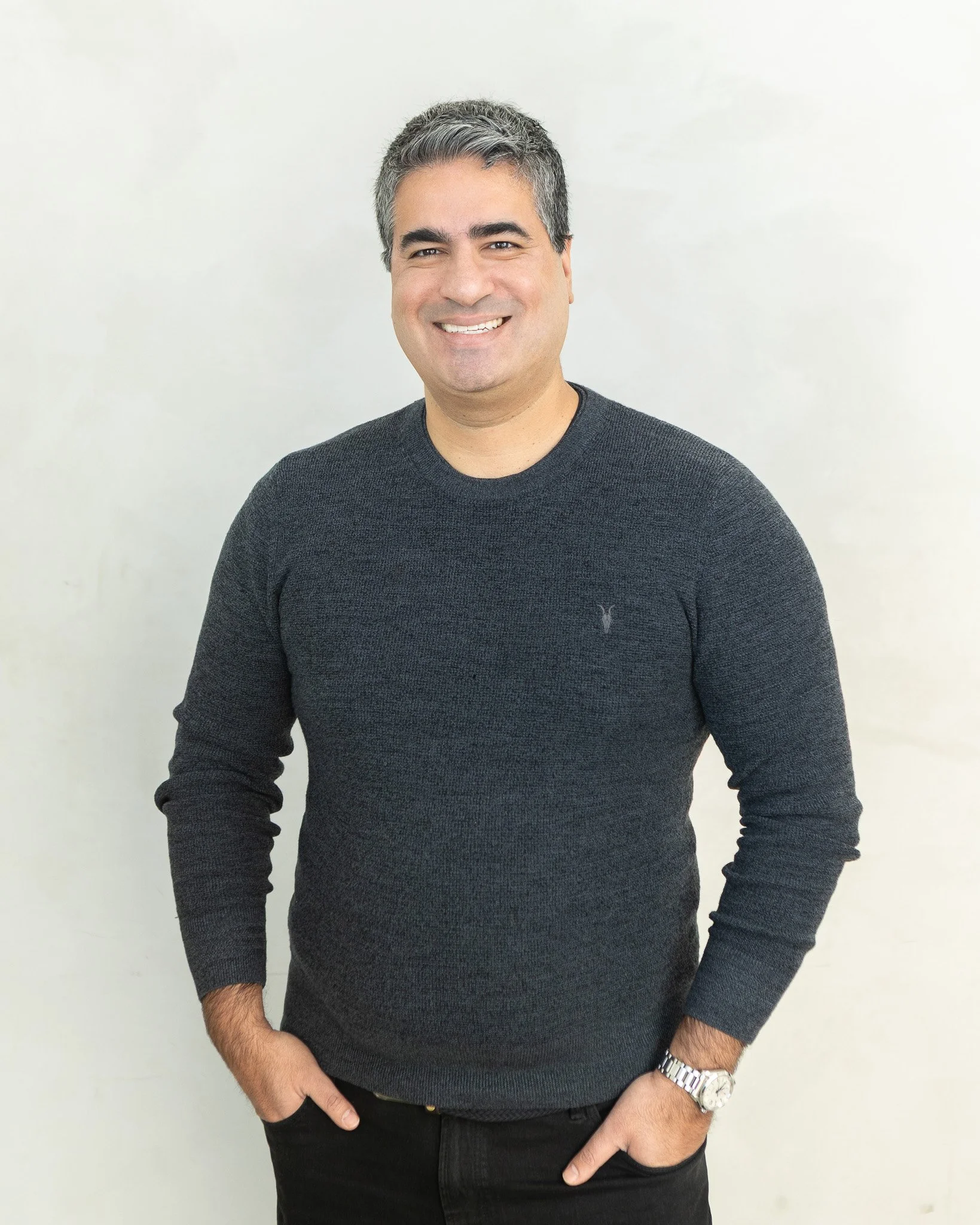 A man with gray hair and a friendly smile wearing a dark gray sweater, standing against a plain light background, with his left hand in his pocket and wearing a silver watch.