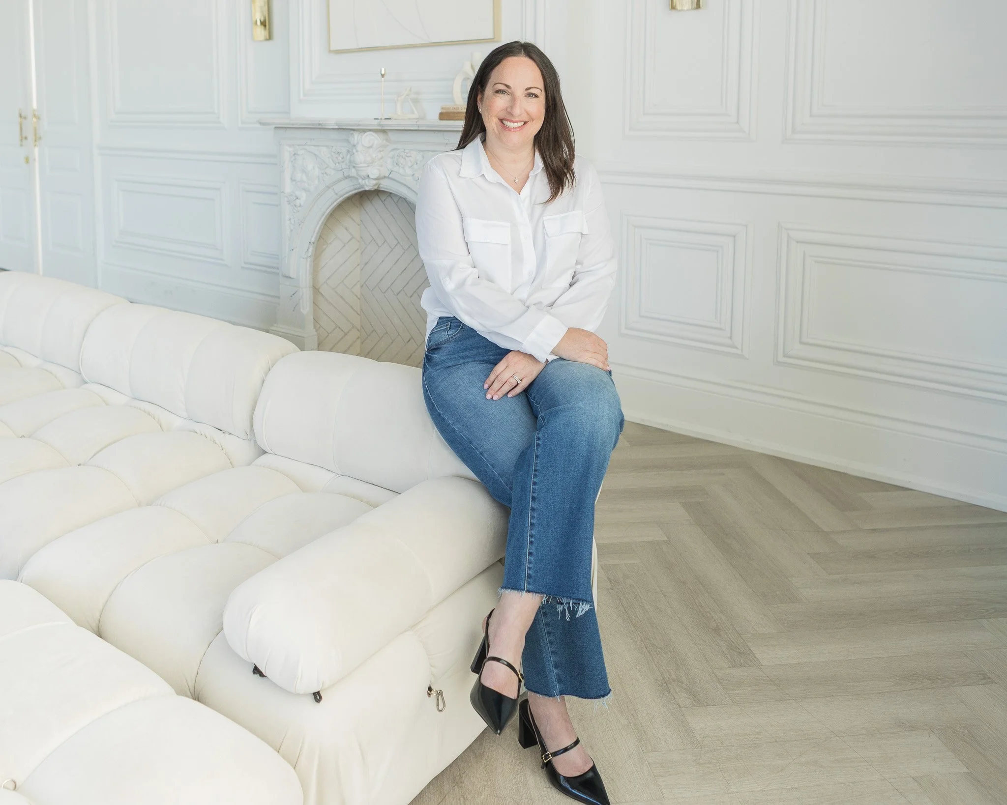 Brunette woman in white shirt seated on coach with marble fireplace in the background
