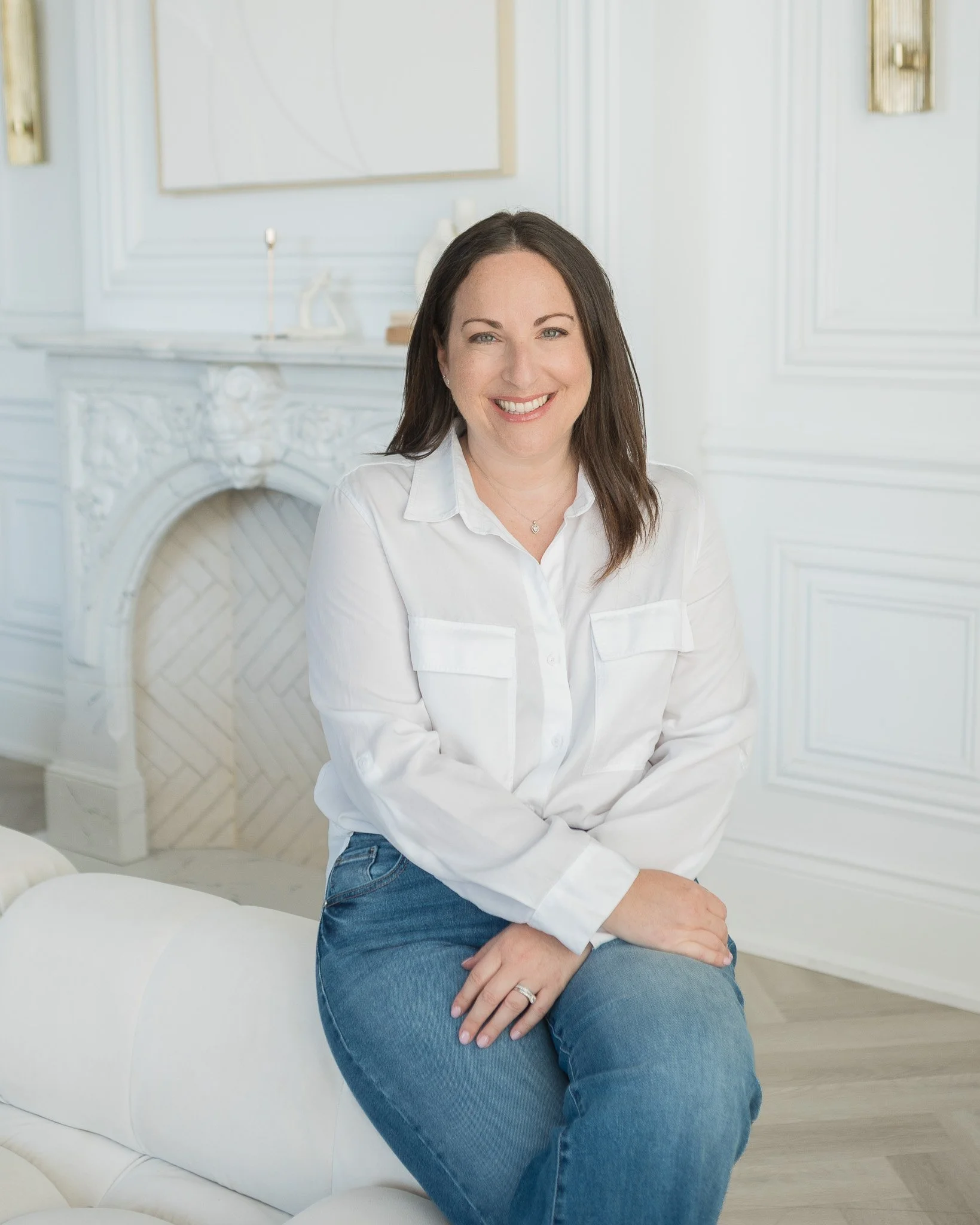 Brunette woman in white shirt seated on coach with marble fireplace in the background