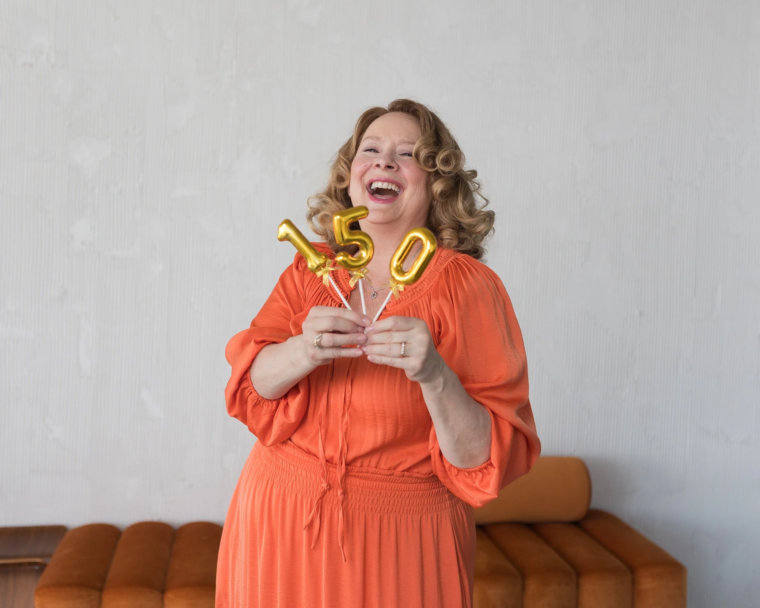 red hair woman in orange dress laughing holding 150 gold sign