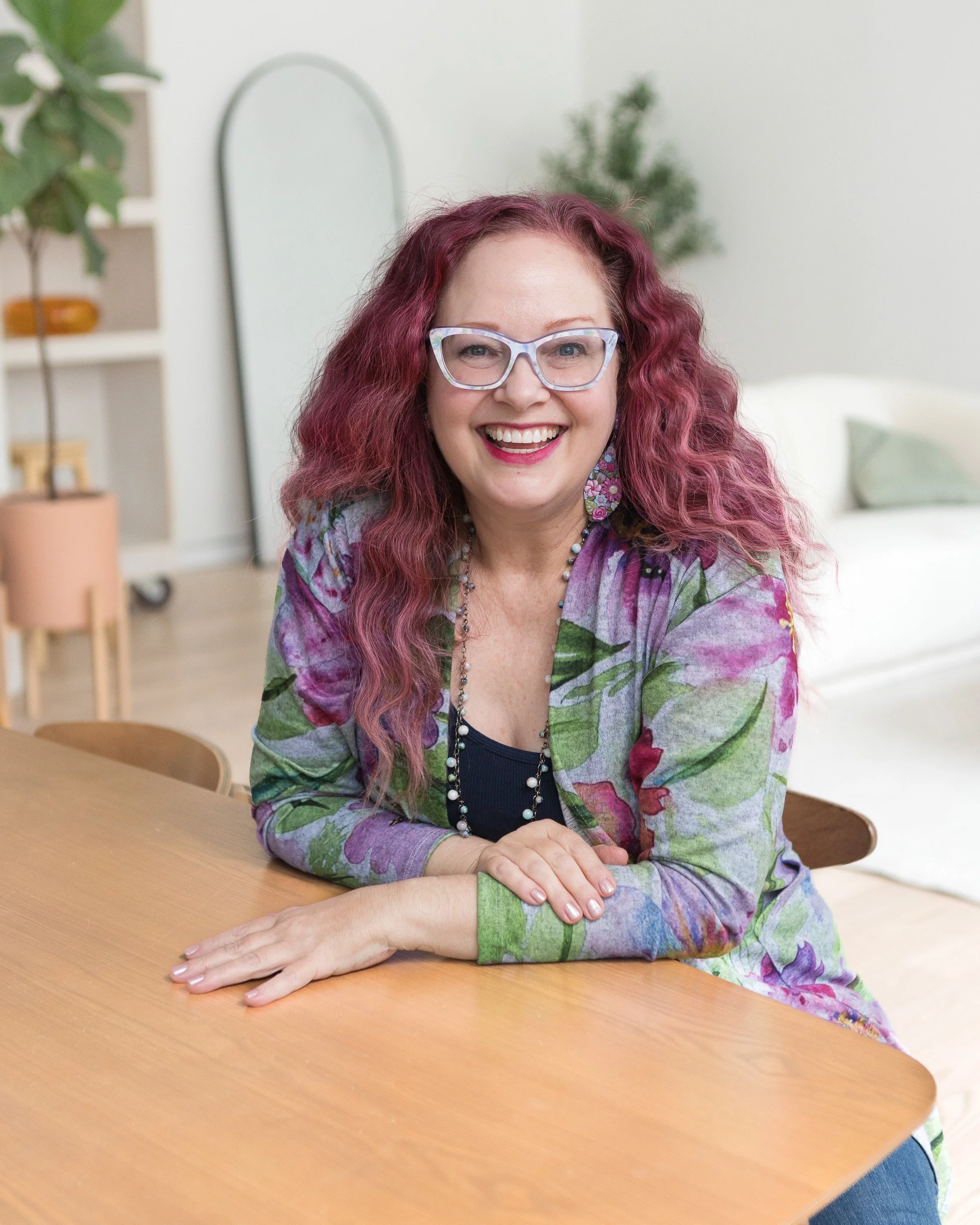 A woman with purple and pink hair and glasses, smiling, seated at a wooden table in a bright room with plants and a mirror in the background.