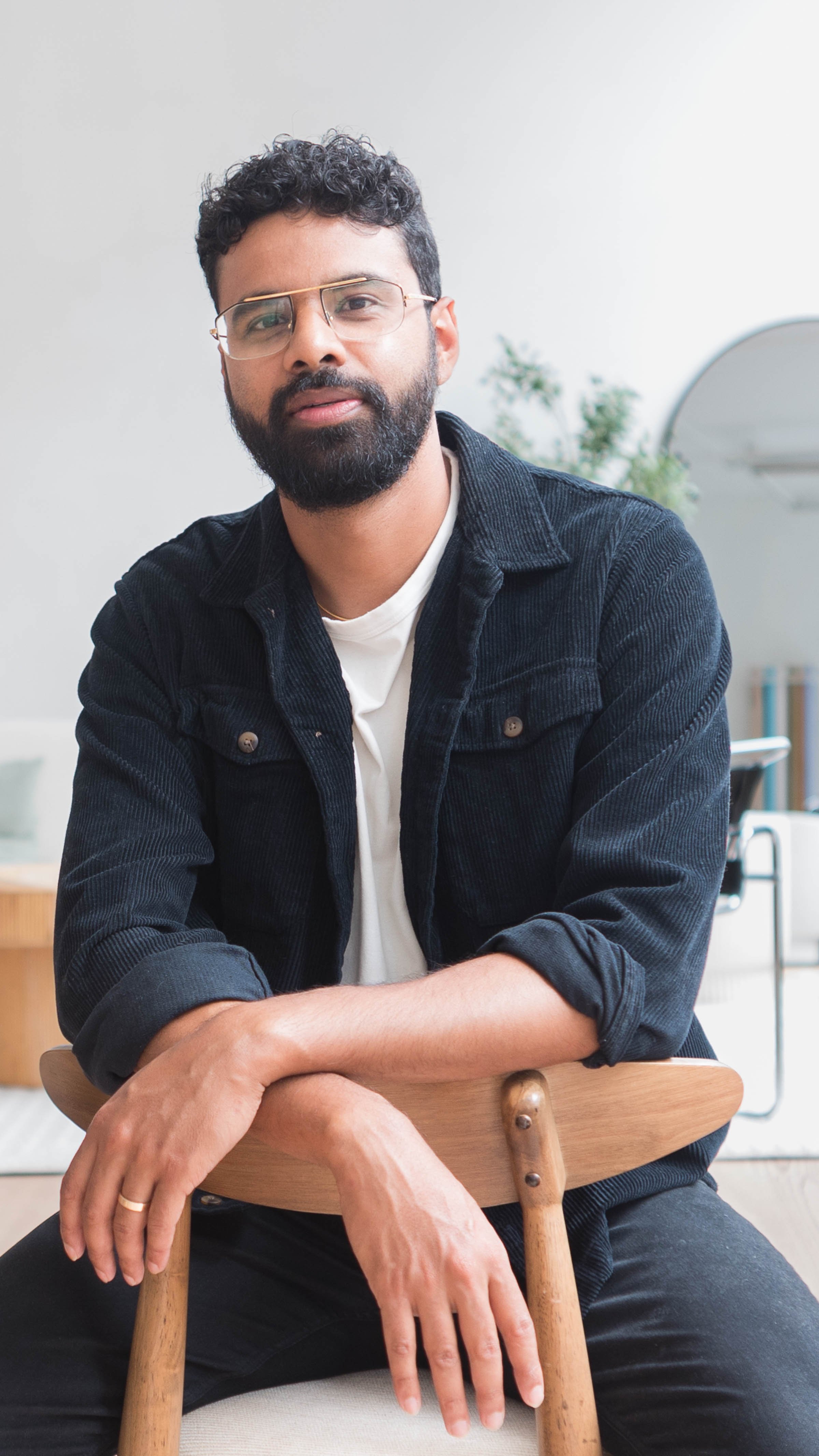 Young man with curly hair, glasses, and a beard sitting on a wooden chair in a modern, minimalistic room with plants and a mirror in the background.